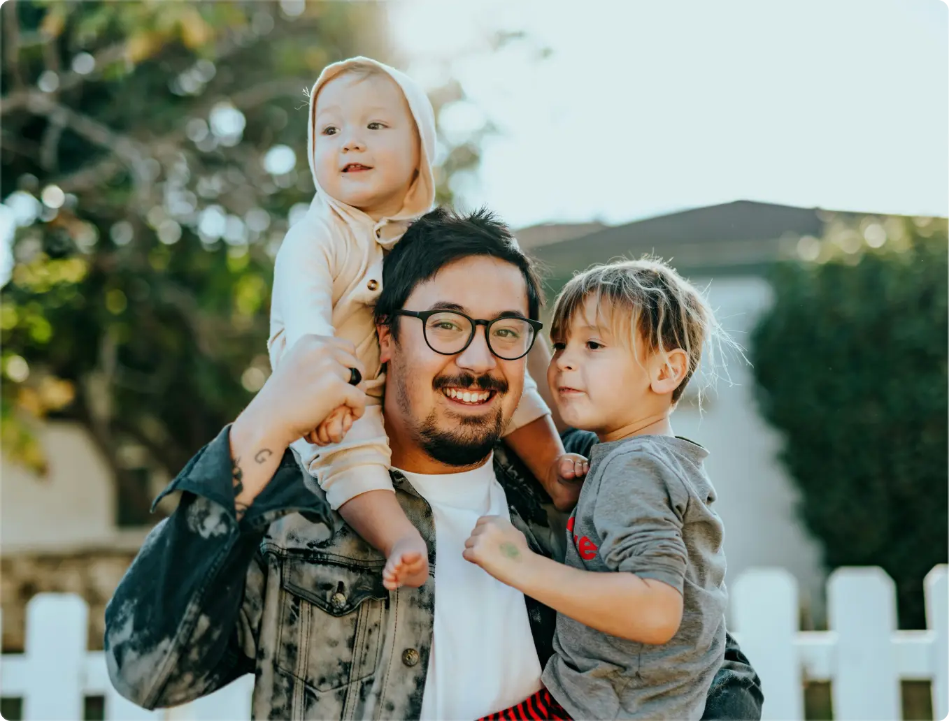 Smiling man wearing glasses holding two young children, one on his shoulders and one in his arms, outdoors near a white picket fence.