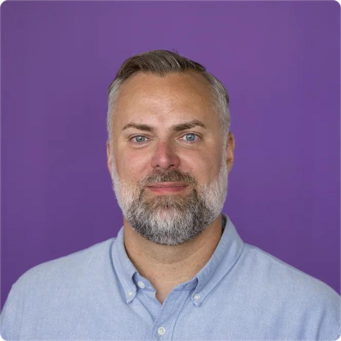 Smiling middle-aged man with gray beard and blue eyes wearing a light blue button-up shirt against a solid purple background.