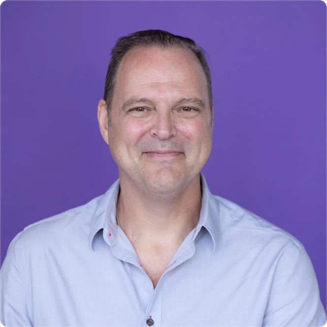 Smiling middle-aged man with short dark hair wearing a light blue shirt against a purple background.