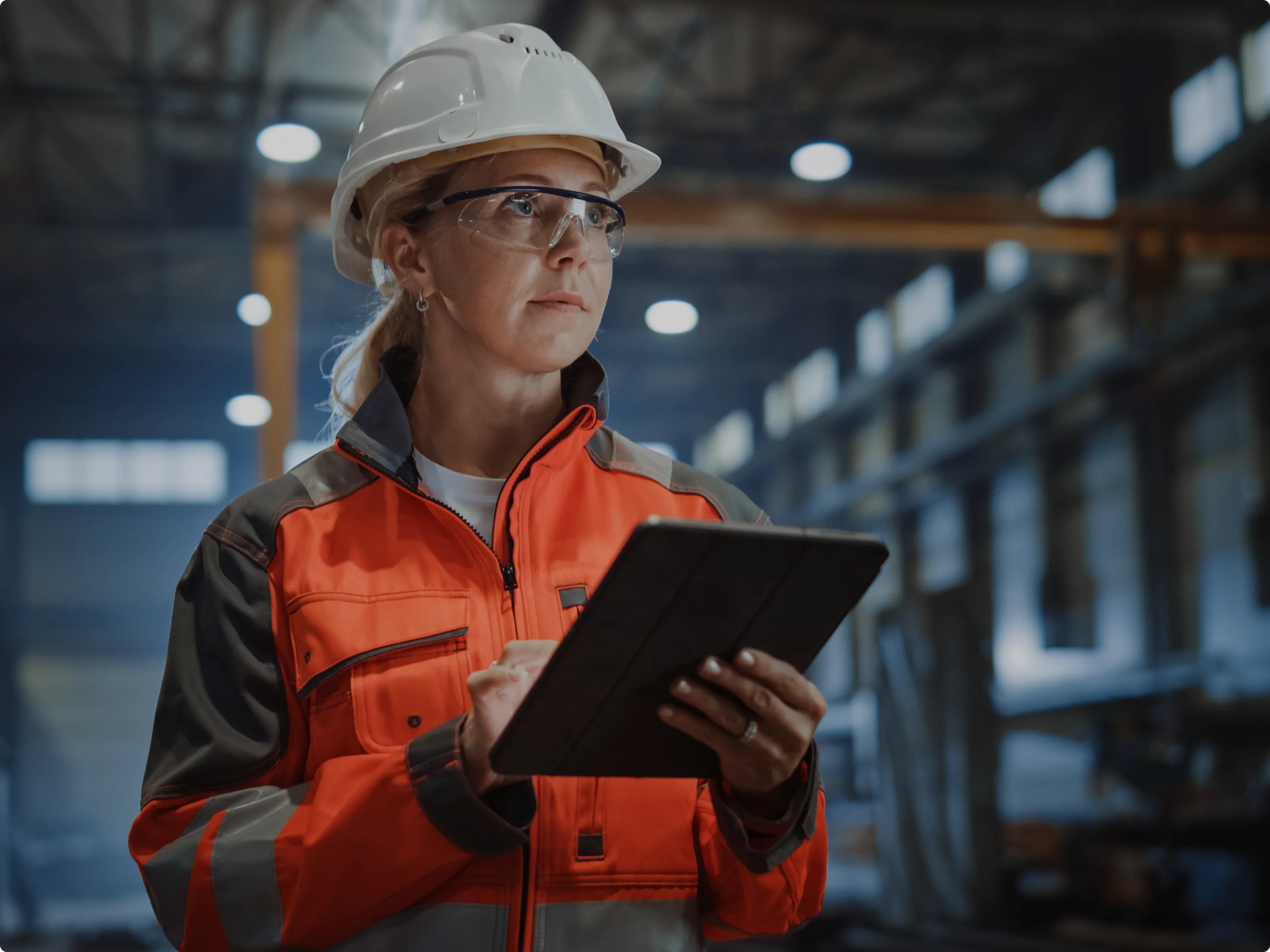 Female industrial worker wearing a white hard hat and orange safety jacket while using a tablet inside a factory.