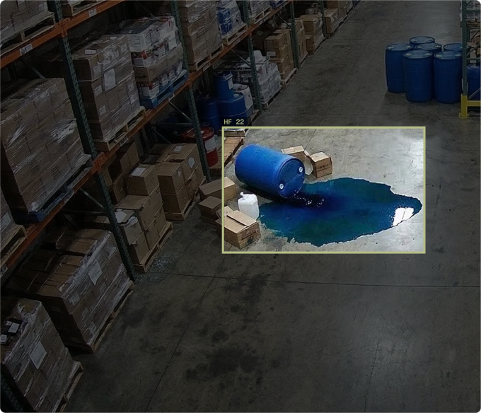 Overhead warehouse camera view of a toppled blue drum leaking a large pool of blue liquid onto the concrete floor, highlighted by a yellow detection box labeled HF 22 to illustrate environmental spill monitoring.