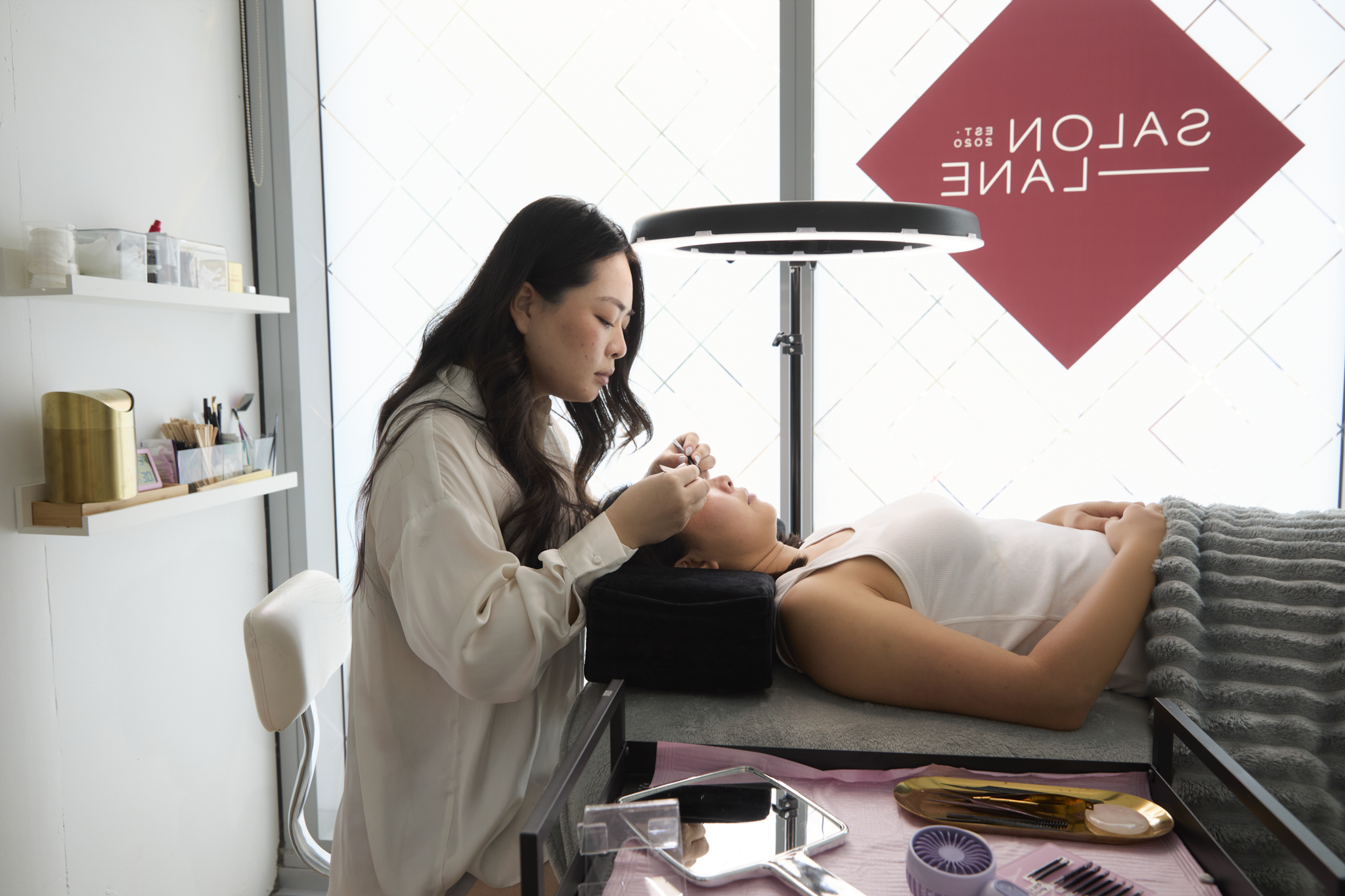Beauty technician applying eyelash extensions to a woman lying on a treatment bed in a salon.