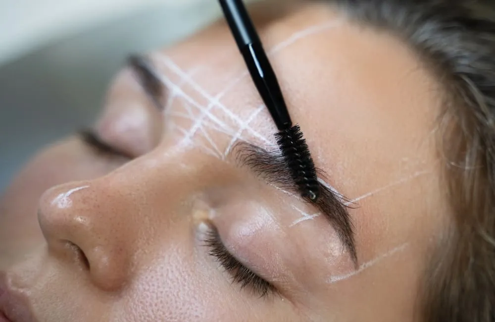 Close-up of a person's face with white guideline markings on the forehead, while a brush combs the eyebrow.