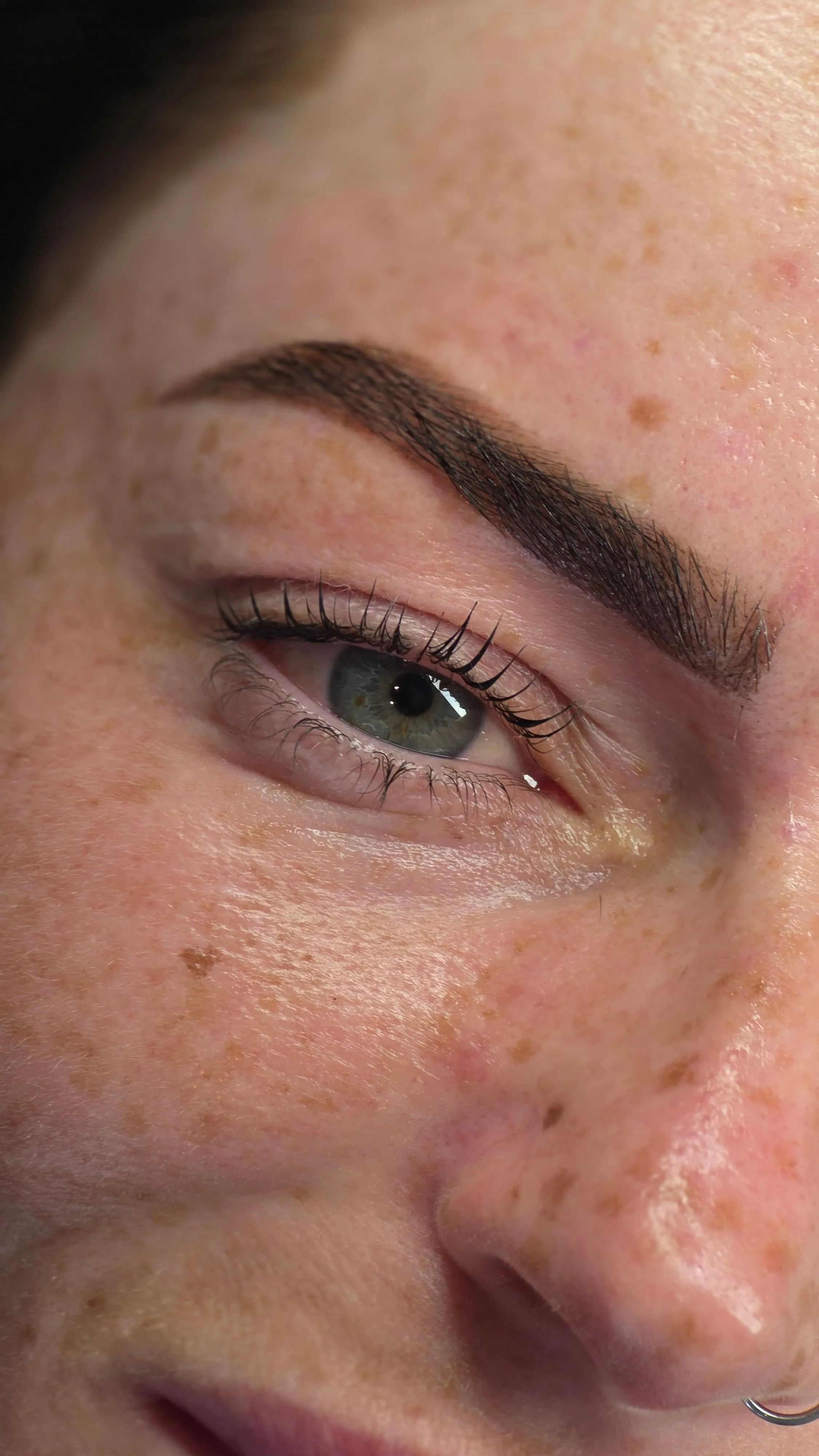 Close-up of a green eye with long lashes on freckled skin and a nose ring.