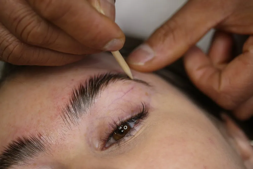 Close-up of a person’s eyebrow being outlined with a wooden stick by another person's fingers.