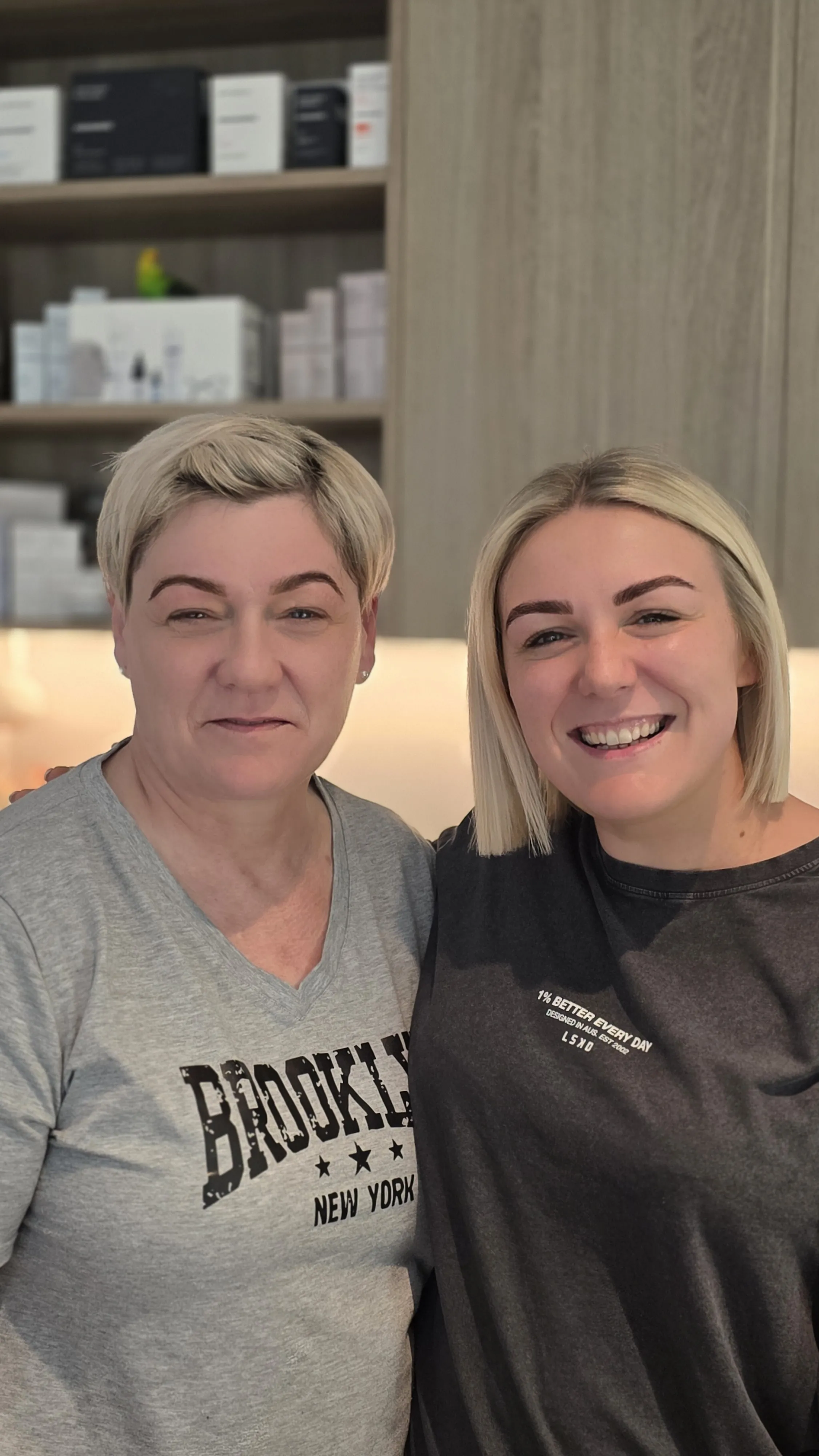 Two women with short blonde hair smiling and standing close together indoors in front of shelves with various boxes.