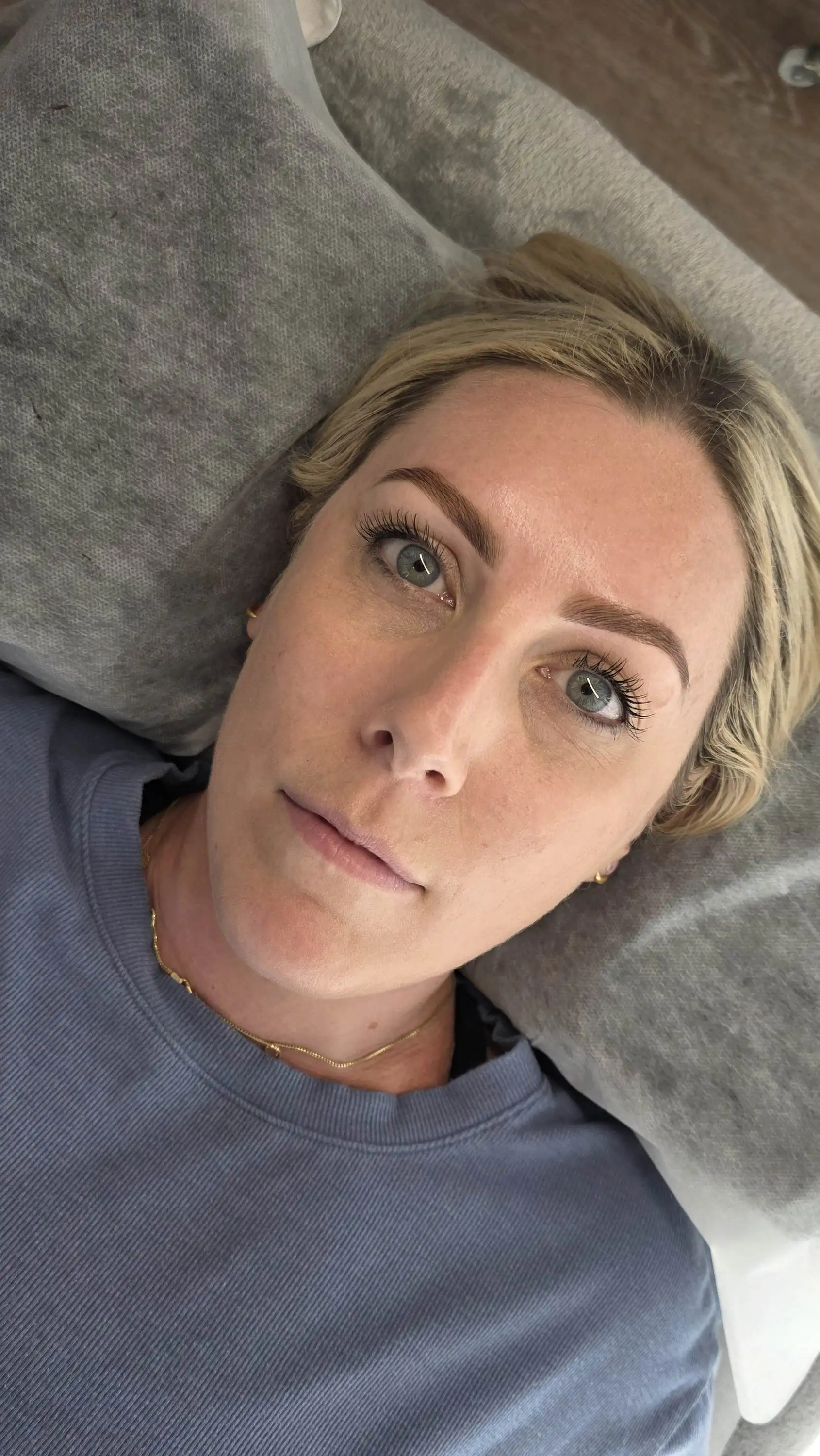 Close-up of a woman with blue eyes and blonde hair lying on a gray pillow wearing a blue shirt.