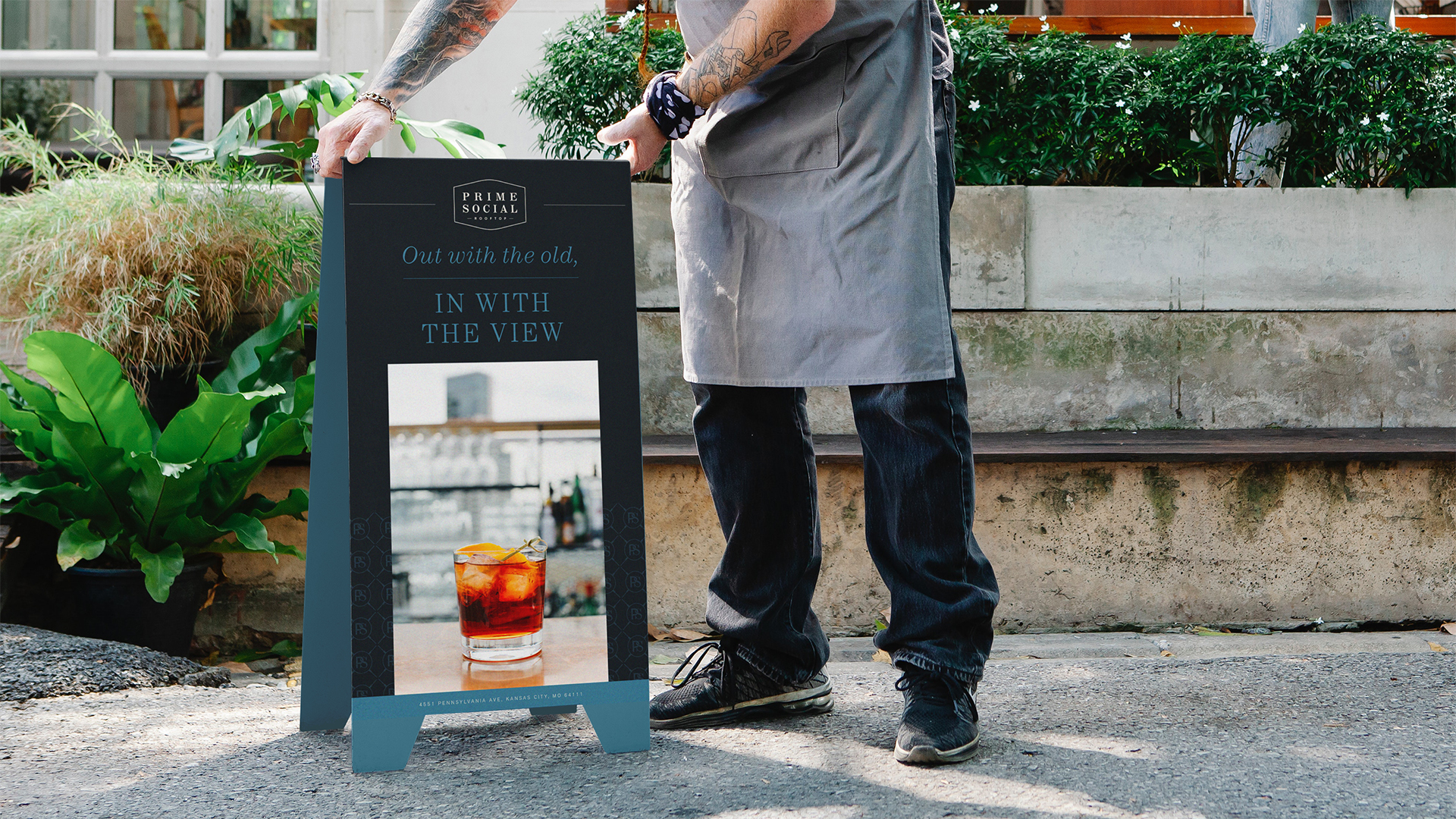 Person in apron holding a Prime Social rooftop sign that features a cocktail with ice and an orange twist on a table.