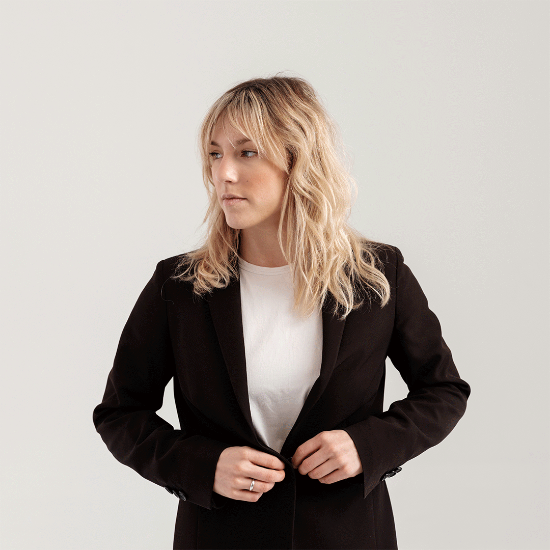 Blonde woman with wavy hair wearing a white shirt and black blazer, buttoning the blazer while looking to the side.
