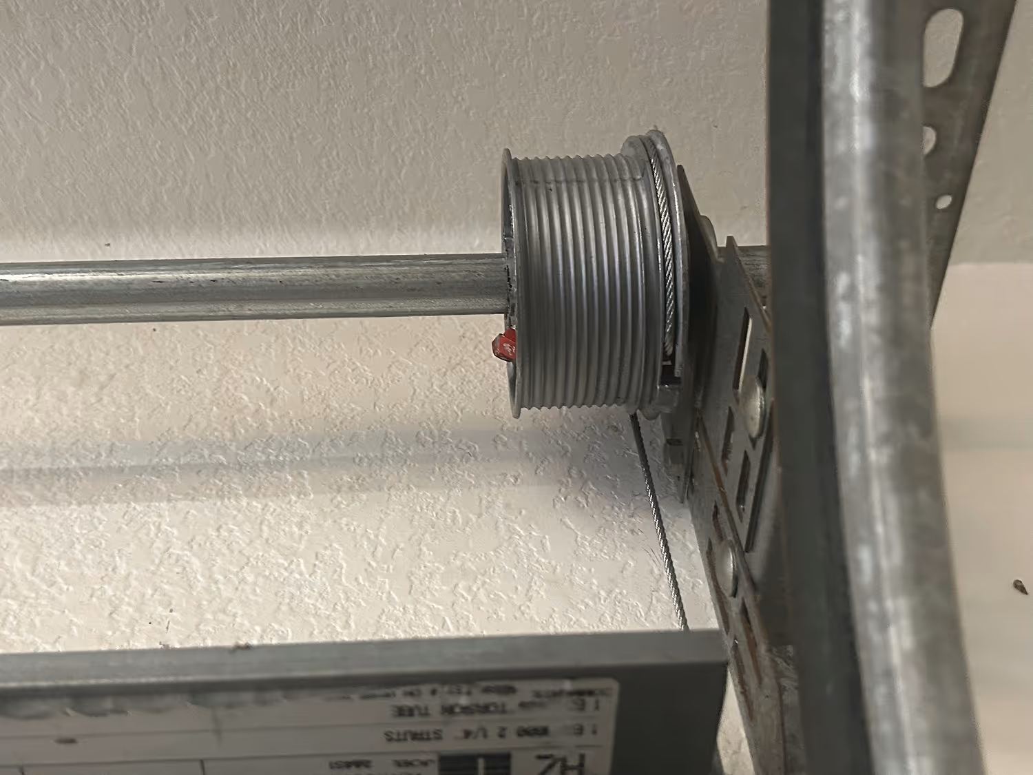 Close-up of a metal garage door torsion spring drum with a cable wrapped around it, mounted on a wall.