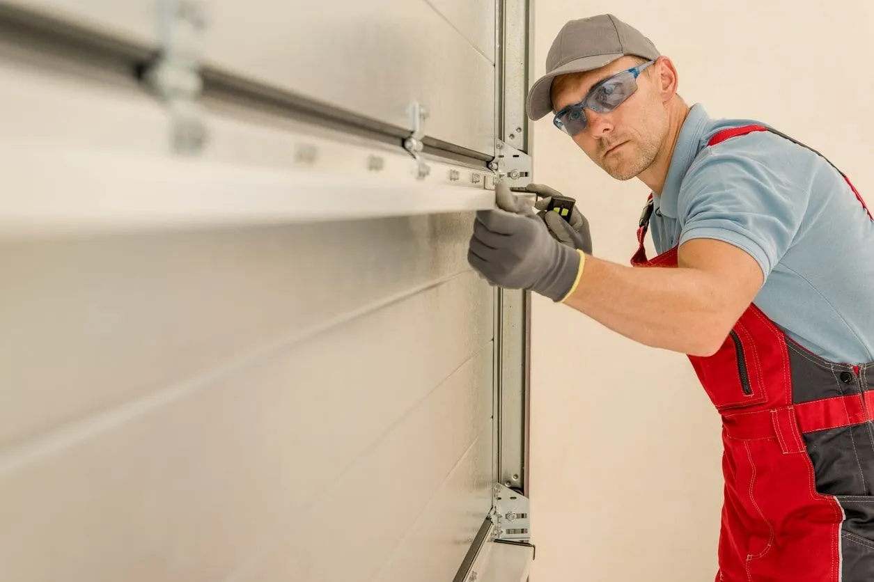 Worker in red overalls and safety glasses adjusting a garage door mechanism.