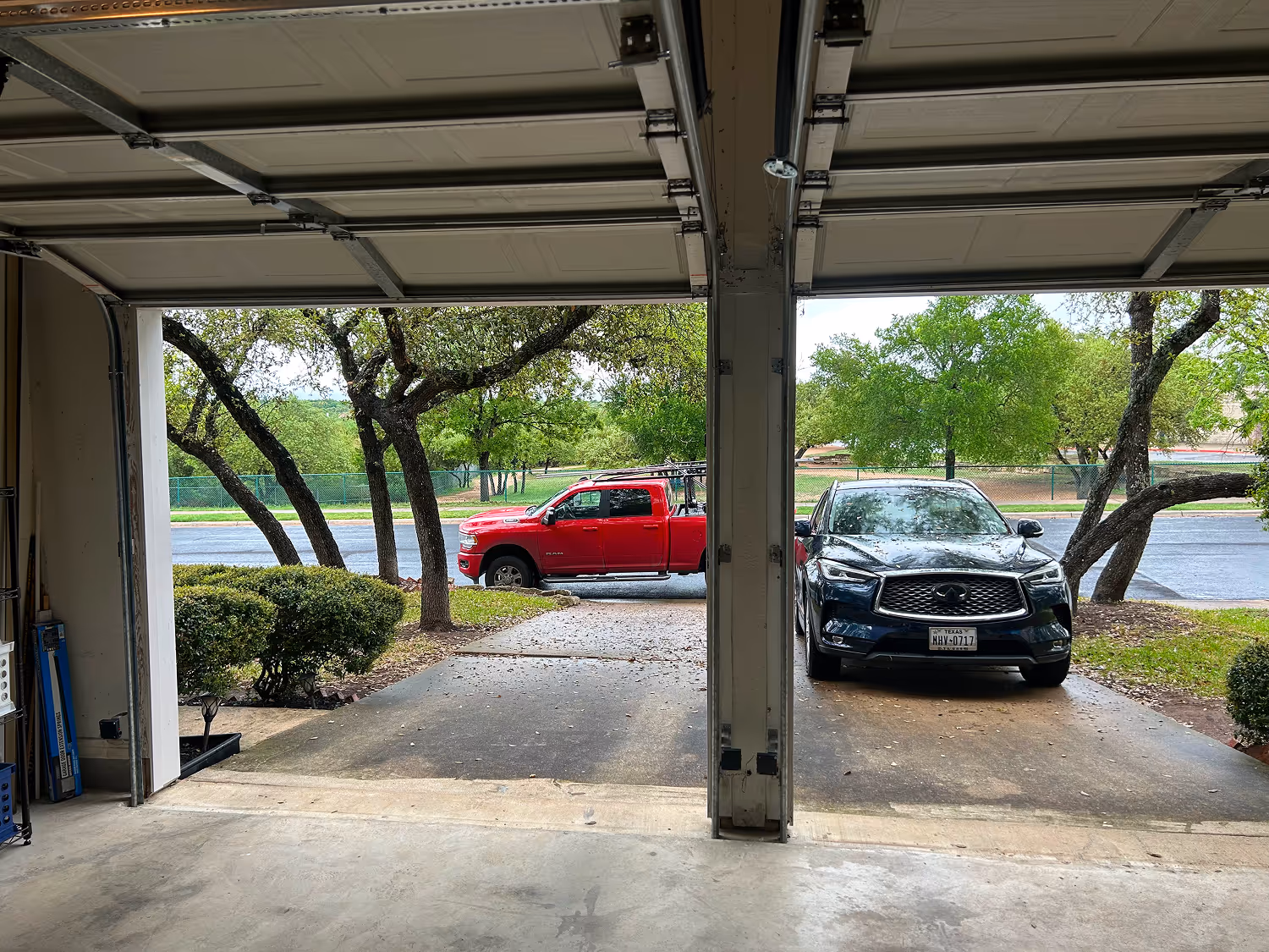 View from inside a double garage with both doors open, showing a red pickup truck parked outside on the left and a black SUV parked on the right driveway, with green trees and bushes around.