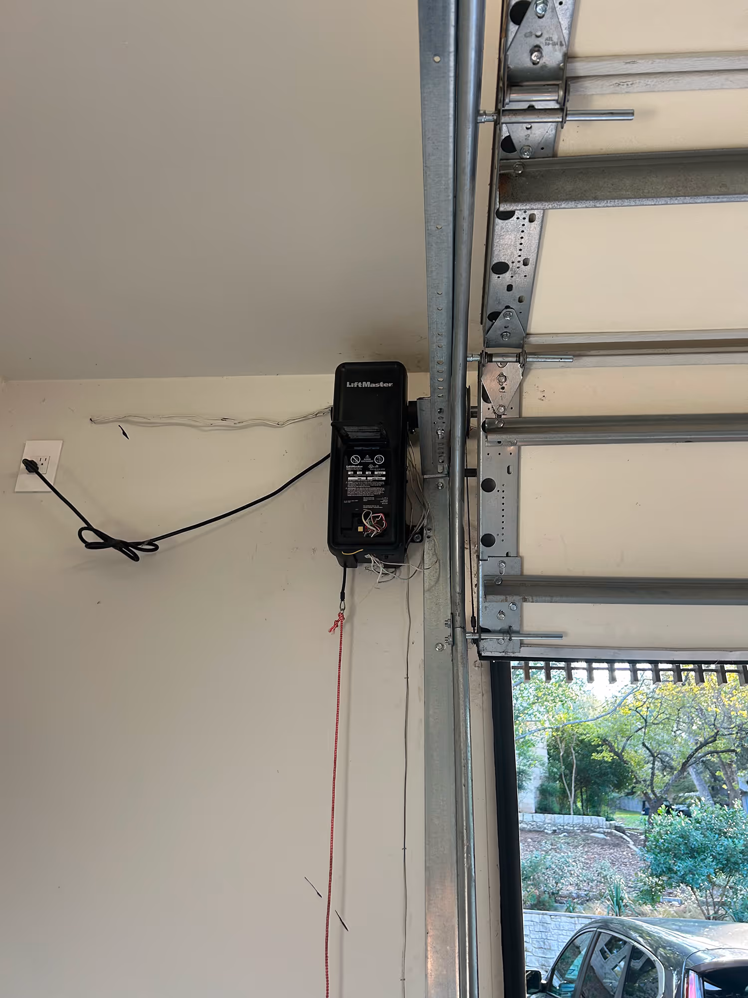 LiftMaster garage door opener mounted on a wall beside a partially open garage door with an outdoor view of trees and a parked car.