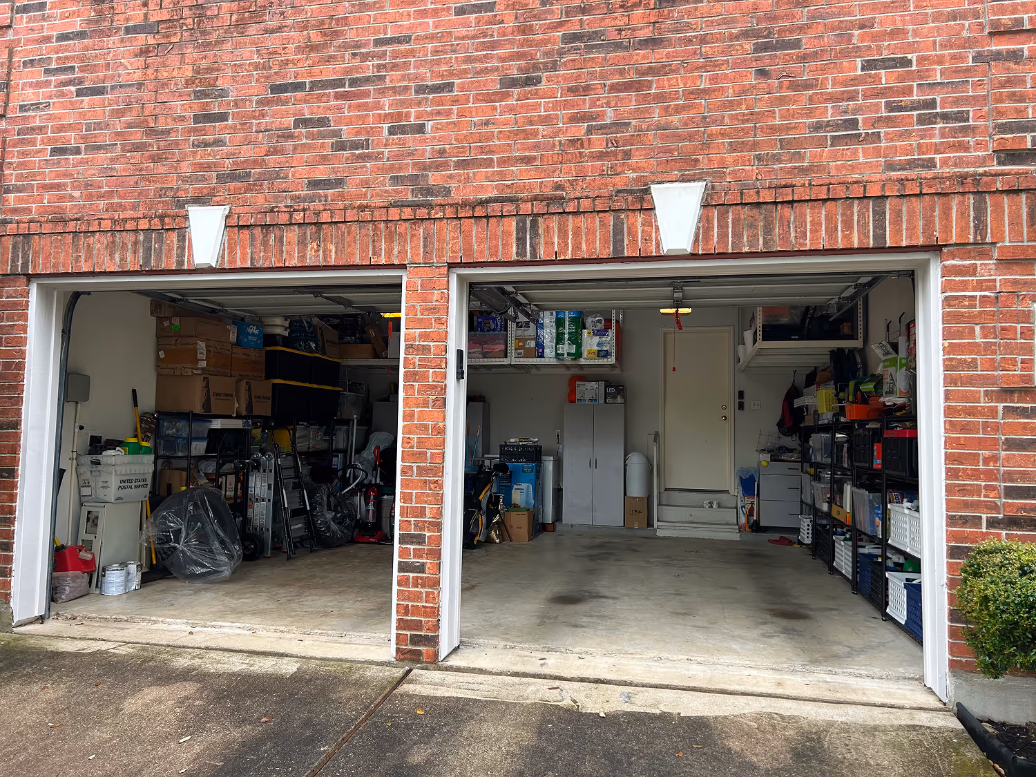 Two-car brick garage with open doors; left side cluttered with boxes and storage items, right side mostly clear with shelves and cabinets along the walls.