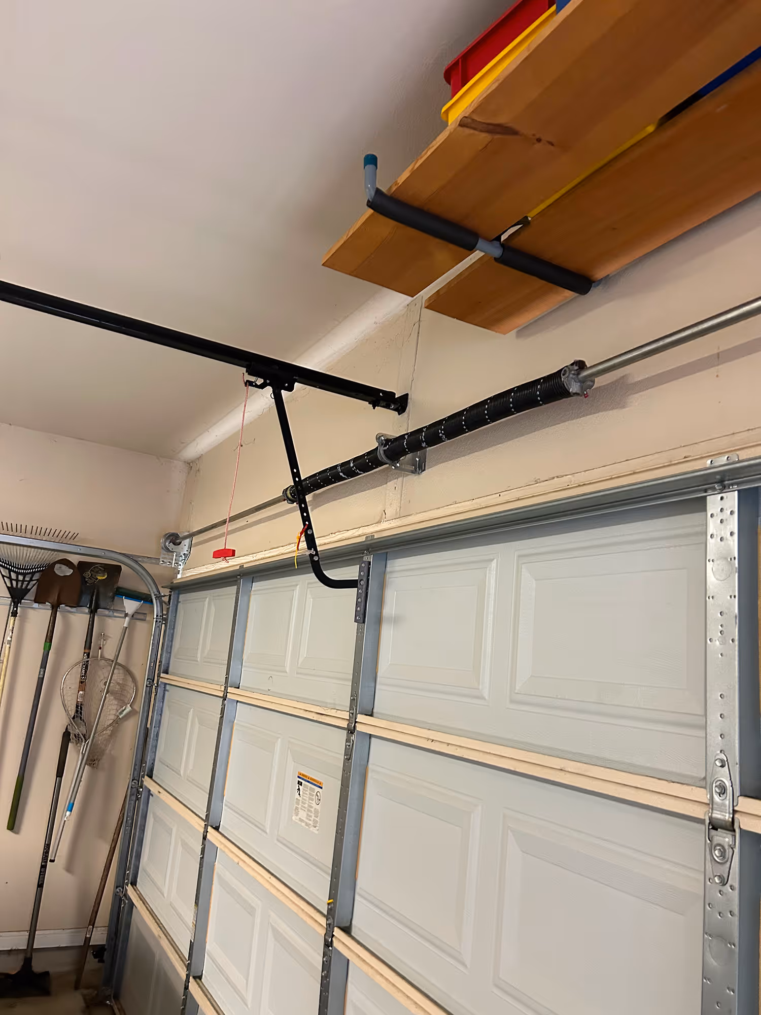 Interior view of a garage door with metal tracks, torsion spring, and a red emergency release cord hanging down; garden tools hanging on the wall and wooden shelves with storage bins above.