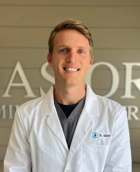 Dr. Andrew Gasior, dentist, wearing a white lab coat over dark scrubs, smiling confidently in a professional portrait against a neutral office wall with soft lighting