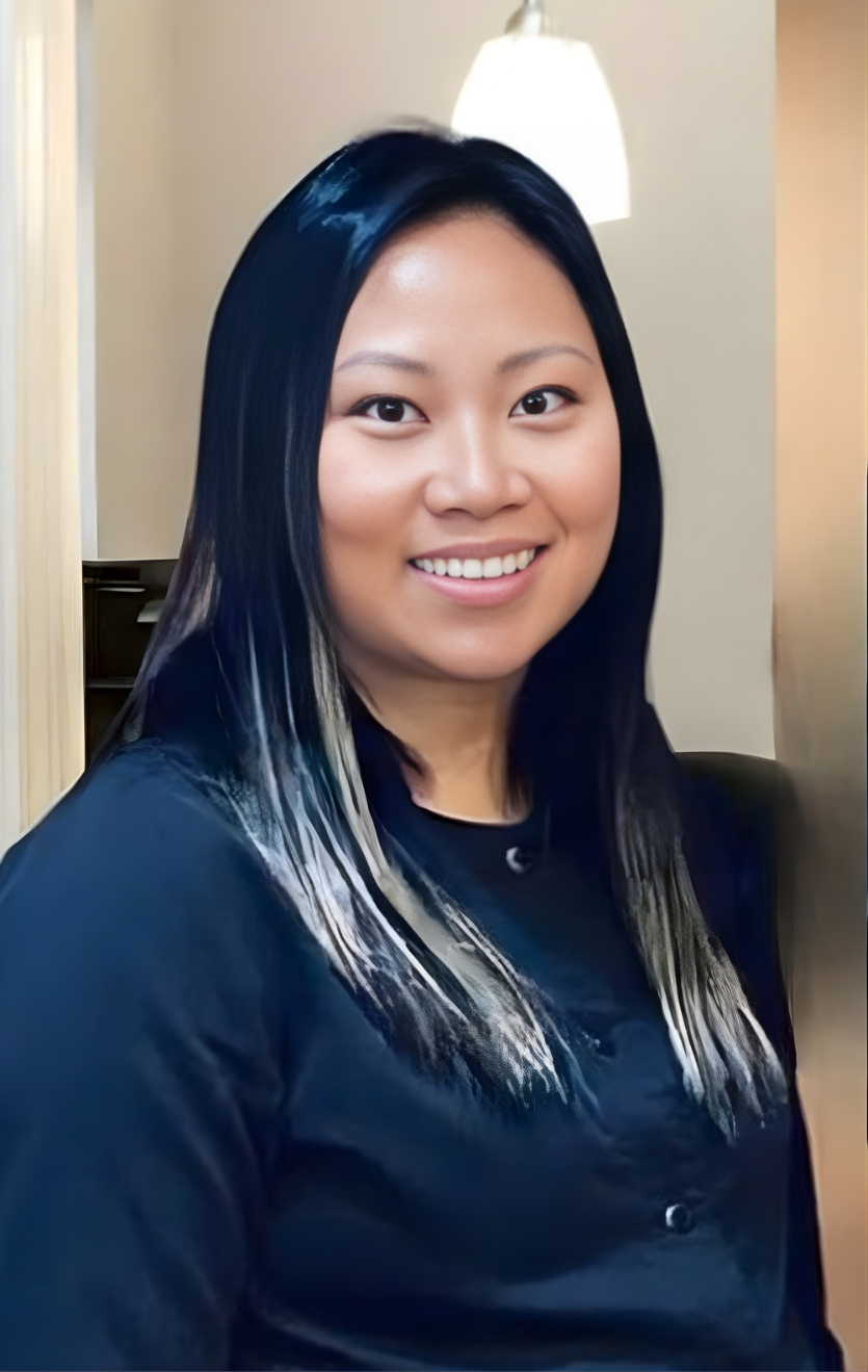 Dr. Angela Khamphouy, dentist, smiling in a professional portrait with long dark hair, wearing a navy blouse, standing in a softly lit office hallway