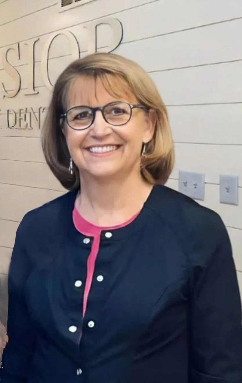 Dr. Maryann Griffith, dentist, smiling warmly in a professional headshot with short light-brown hair and glasses, wearing a dark blouse with a subtle pink accent, standing in front of a textured office wall