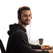 Man wearing headphones and a black hoodie sitting at a desk with a keyboard and a mug.