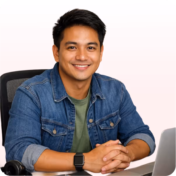 Smiling man wearing a denim jacket seated at a desk with a laptop and headphones.