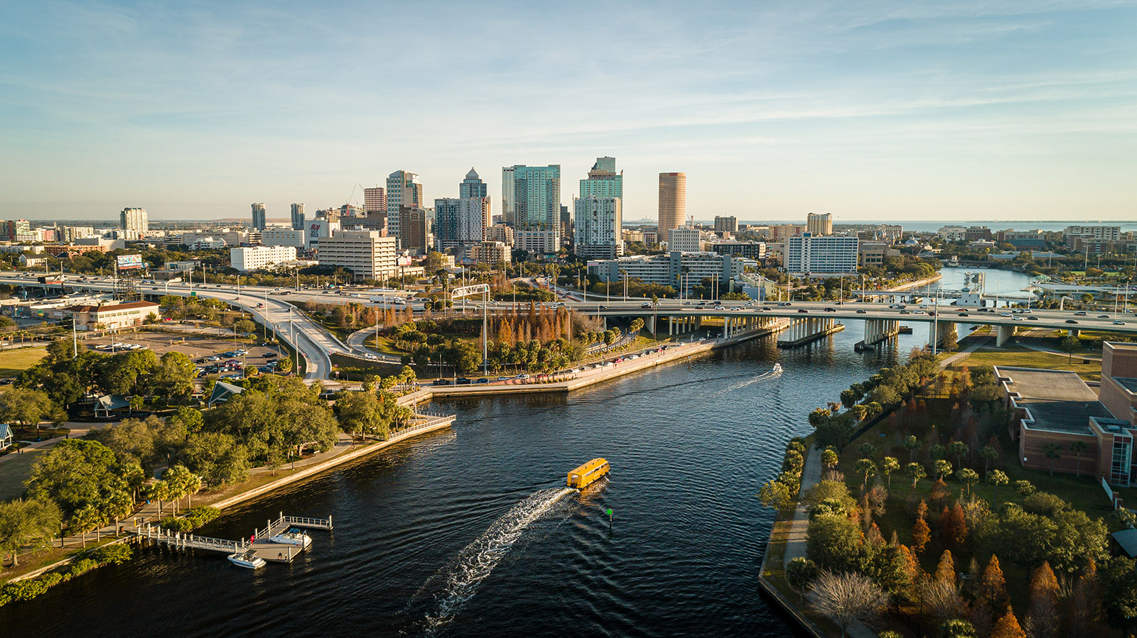 Aerial view of downtown Tampa, Florida skyline along the Hillsborough River, featuring high-rise buildings, multiple bridges with traffic, palm trees, and a yellow boat creating a wake on the water at sunset.