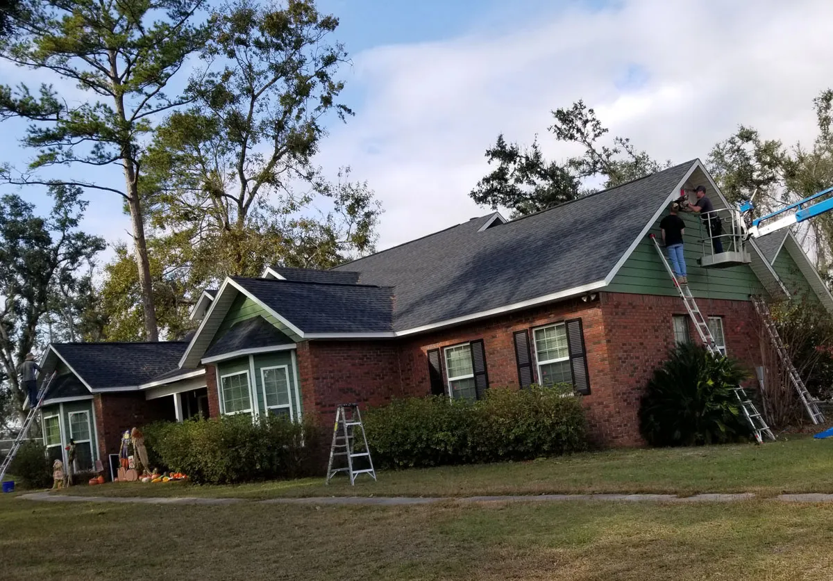 Roof replacement in progress by Florida building contractor on a large brick and green siding residential home with crew on site