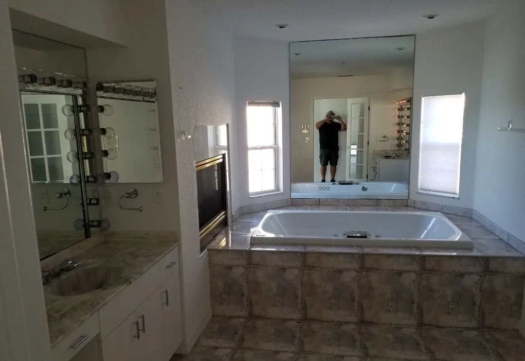 Bathroom with a built-in bathtub surrounded by tiled platform, large mirror, two windows, and a vanity with a sink and lighted mirror.