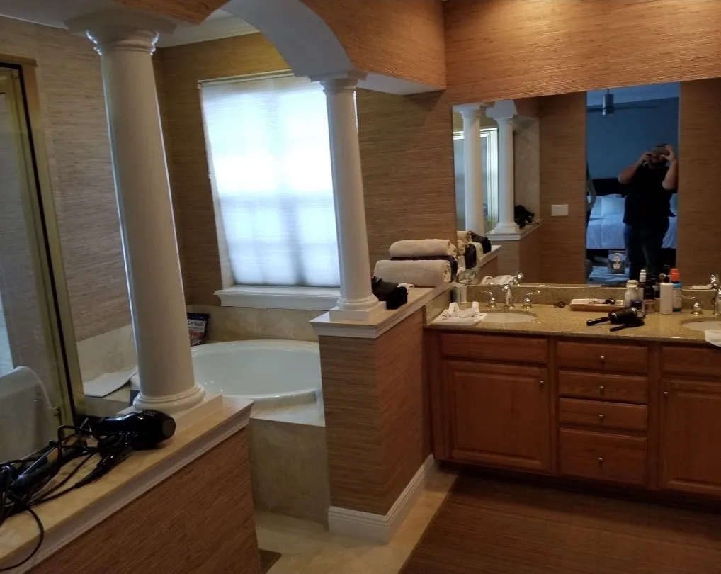 Bathroom with beige walls, a bathtub behind white columns, wooden vanity with dual sinks, towels, and toiletries on the counter.