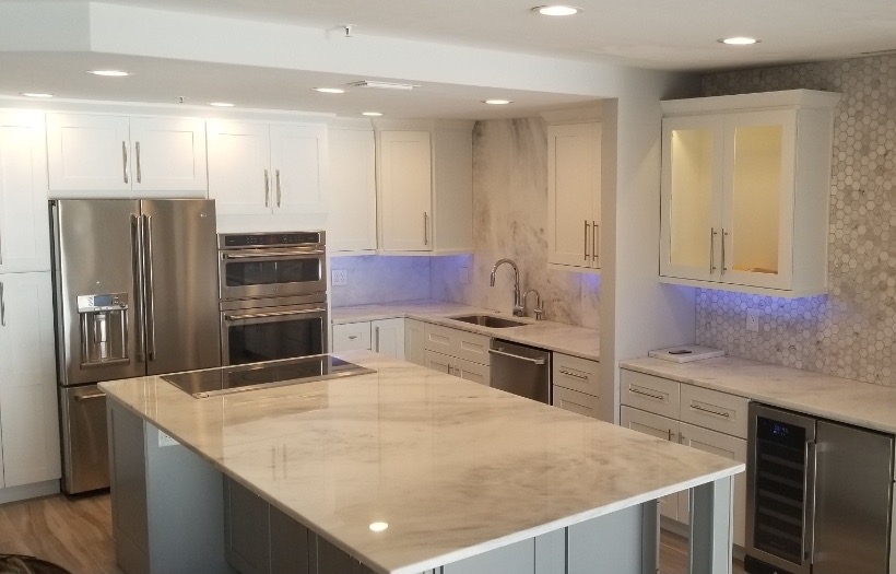 Modern kitchen with white cabinets, stainless steel refrigerator, double oven, large marble island, and subtle blue under-cabinet lighting.