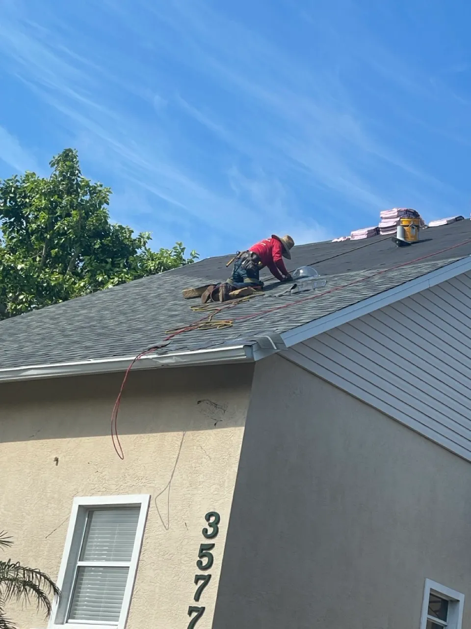 Worker wearing a red shirt and hat kneeling and installing shingles on a house roof under clear blue sky.