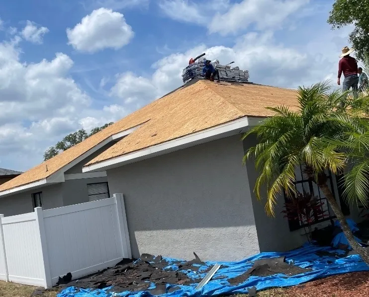 Workers installing new roofing shingles on a house under a partly cloudy sky.