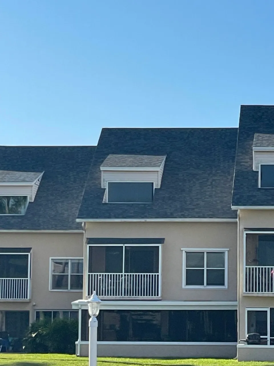 Row of beige townhouse-style buildings with dark shingle roofs and balconies under a clear blue sky.