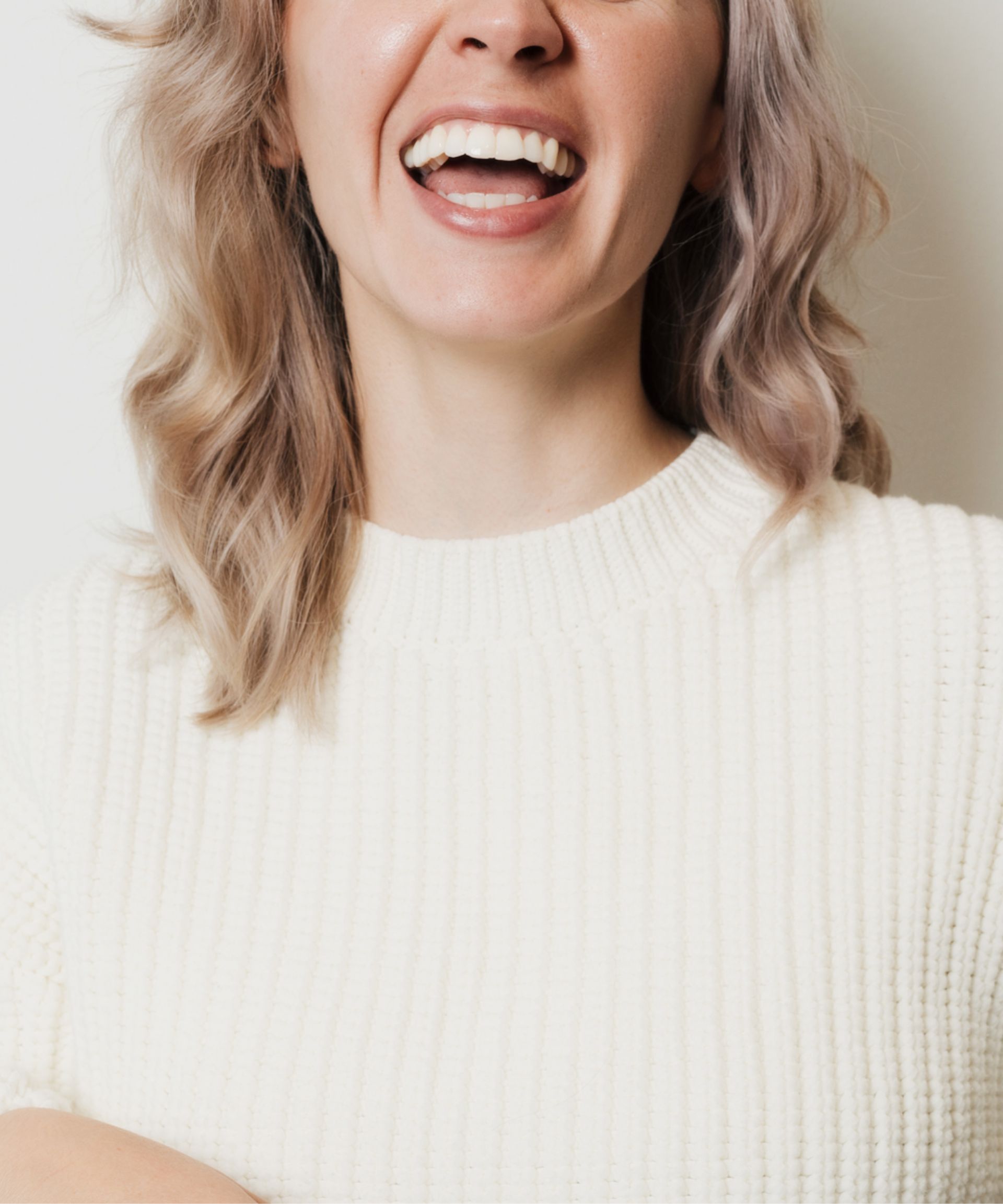 Close-up of a woman with wavy light hair smiling and wearing a white knit sweater.