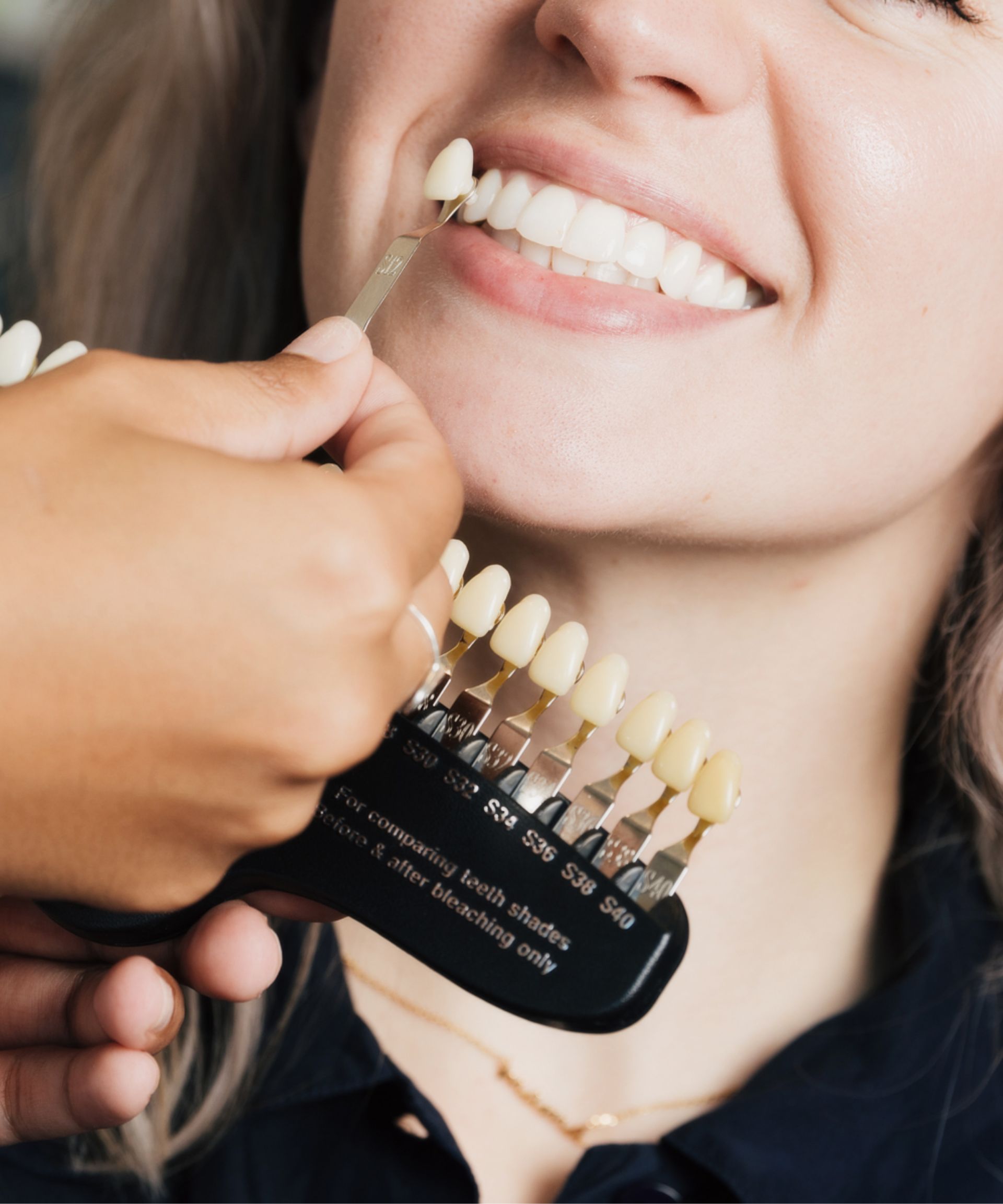 Person smiling as dental professional compares tooth shade guide to their teeth for color matching.