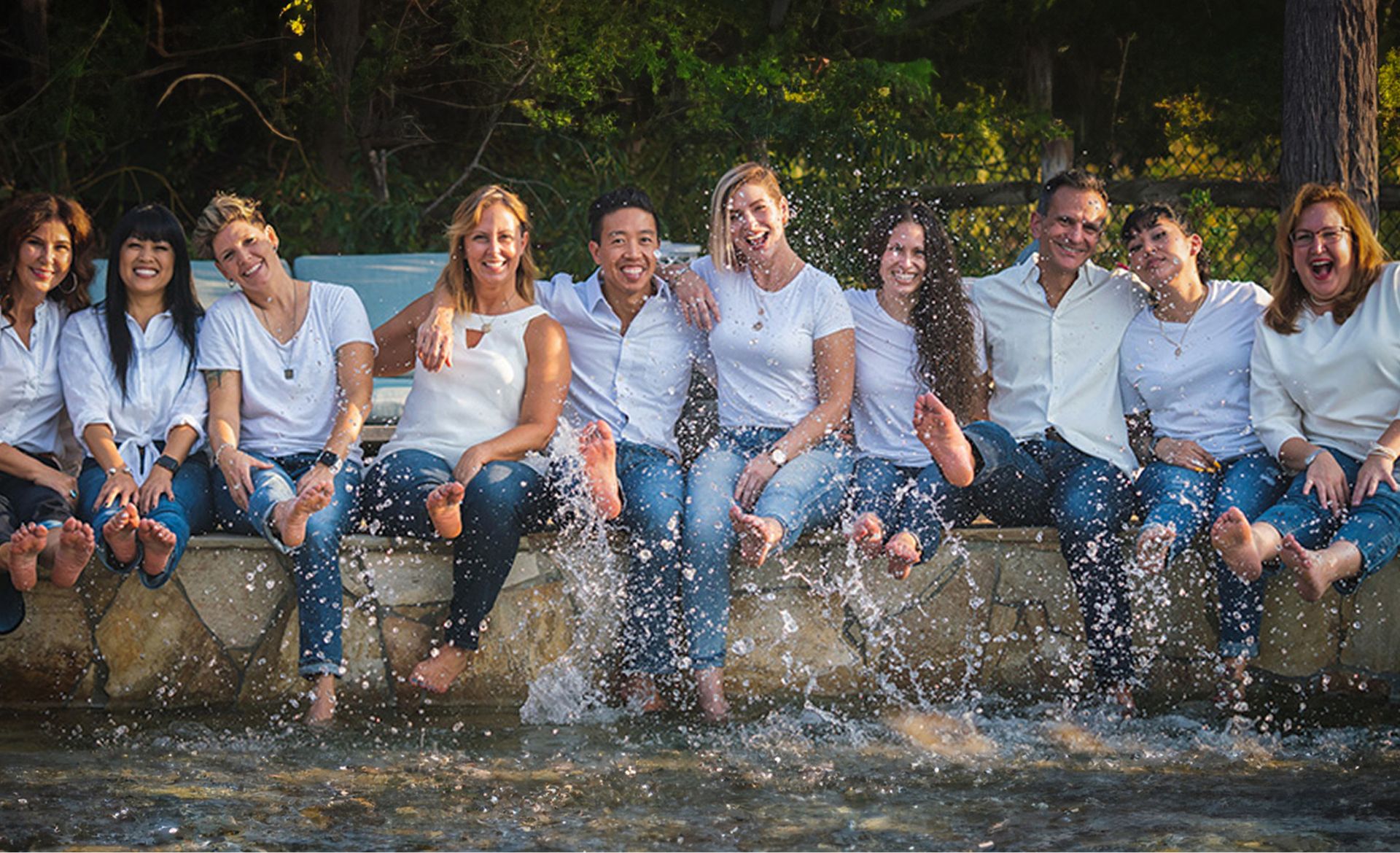 Group of ten people sitting on a stone ledge splashing their feet in water and smiling.