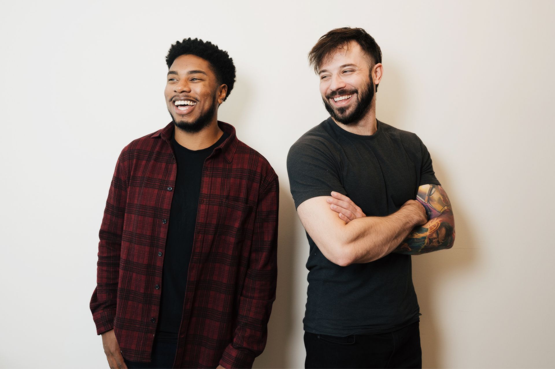 Two smiling men stand side by side, one in a red plaid shirt, the other with tattooed arms crossed.