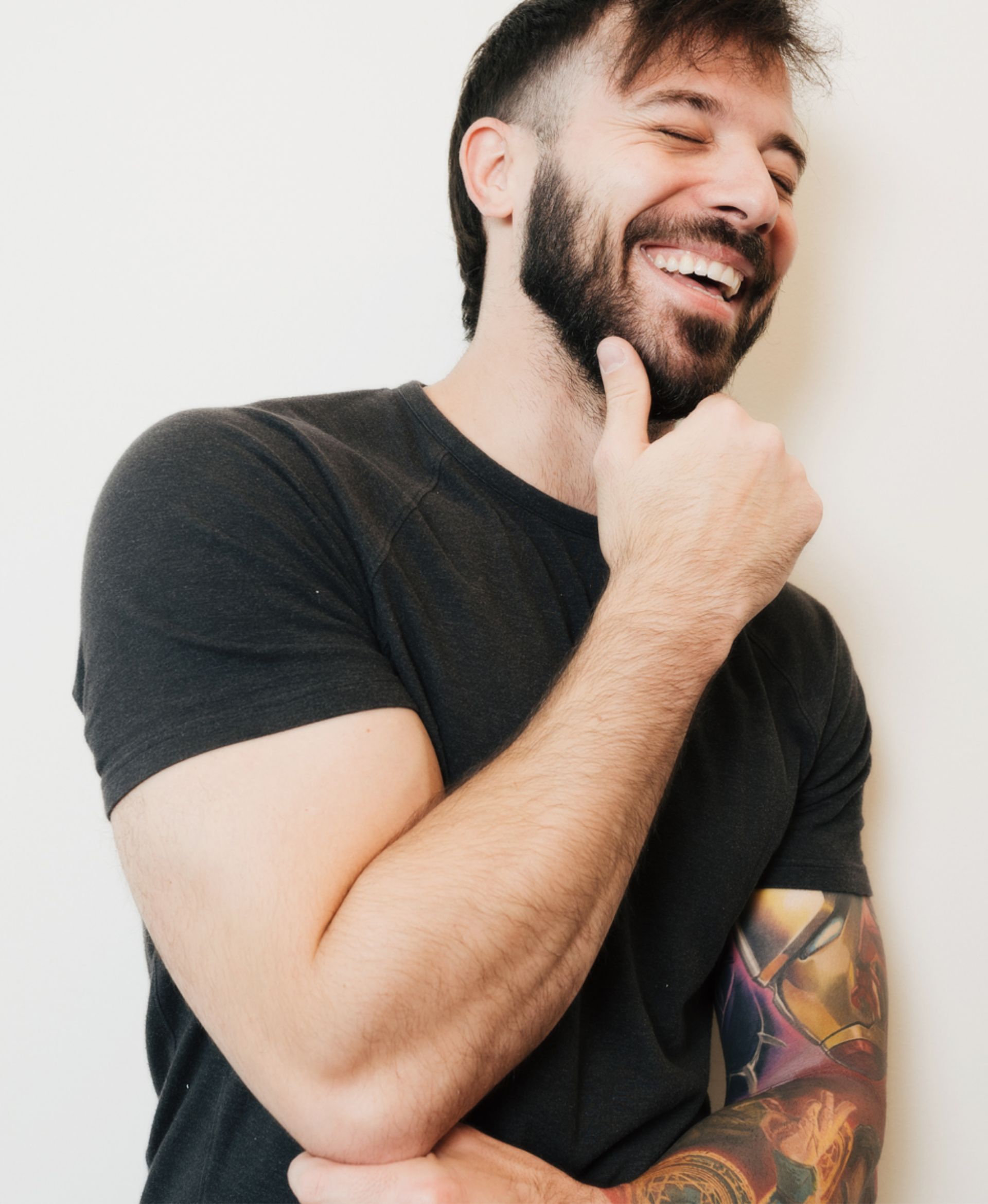 Smiling man with beard, wearing black shirt, showing tattooed right arm, touching chin thoughtfully.