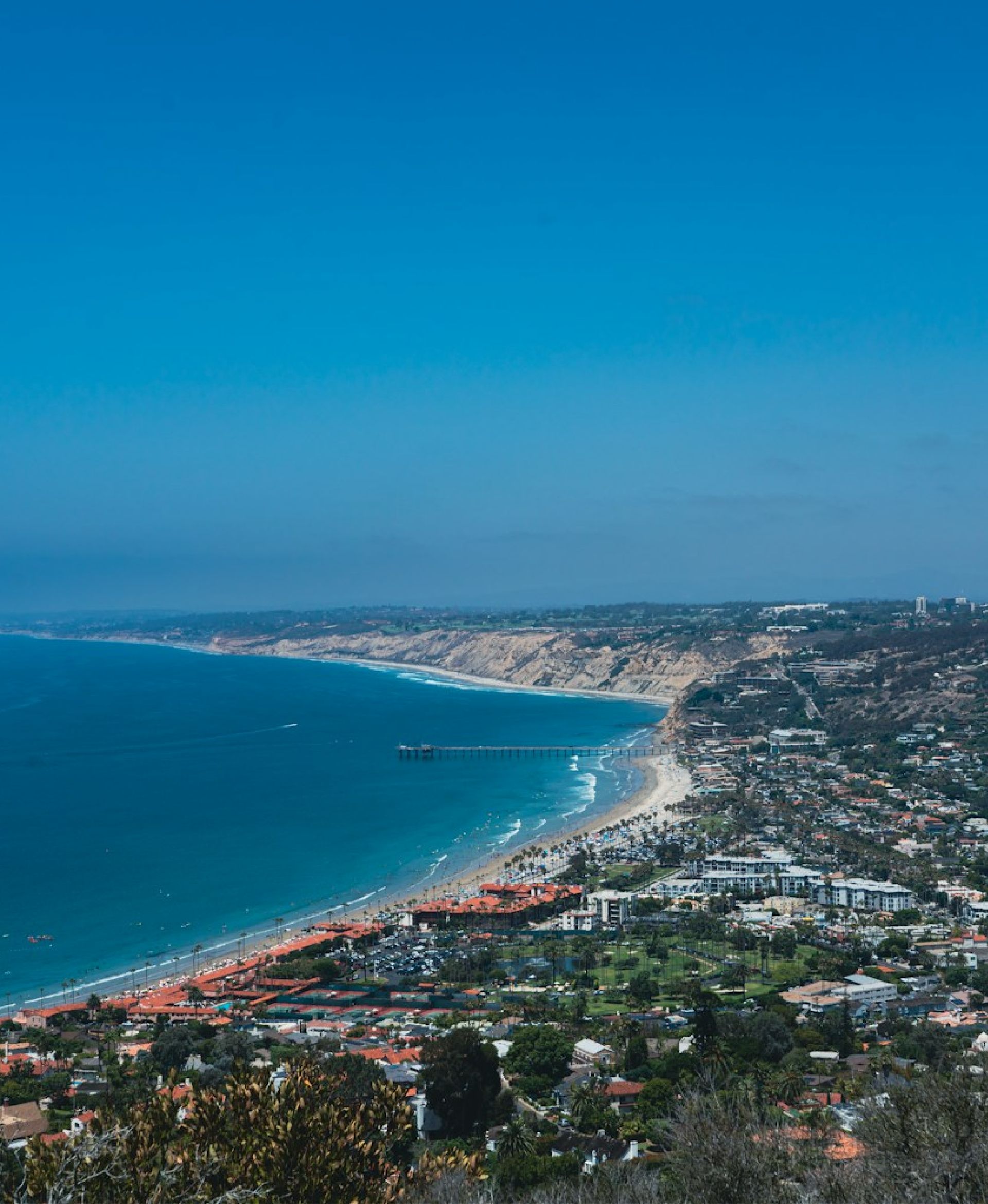 Aerial view of coastal town with beach, pier, ocean, cliffs, and residential buildings under clear blue sky.