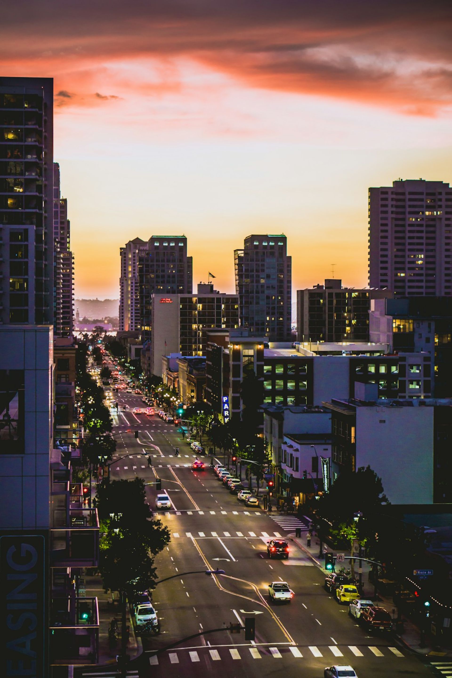 City street with cars, buildings, and trees at sunset with a colorful sky.