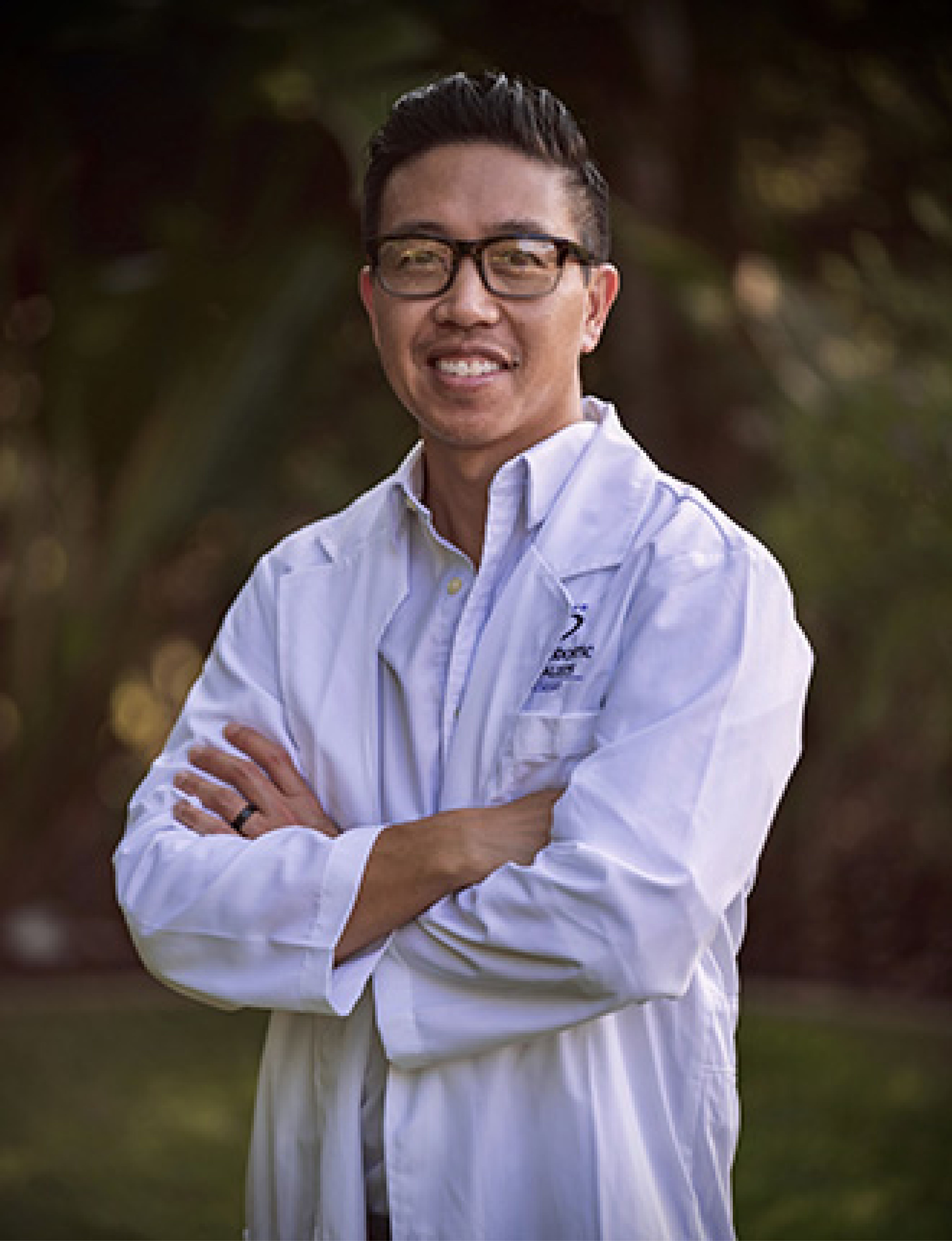 Smiling man wearing glasses and a white lab coat standing outdoors with arms crossed.