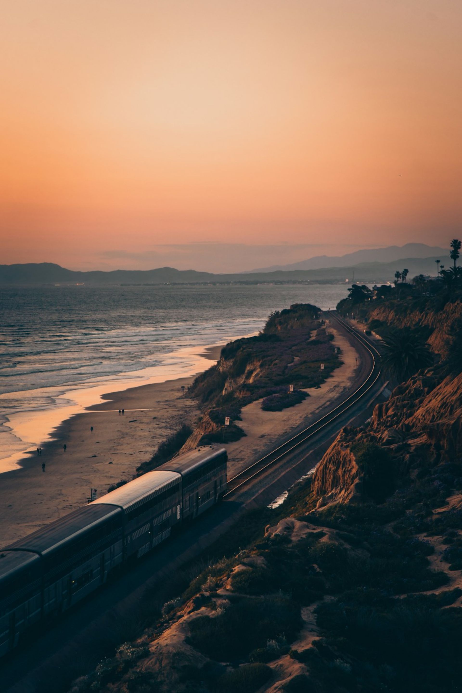 Train traveling along a coastline with beach, ocean waves, and hills at sunset.