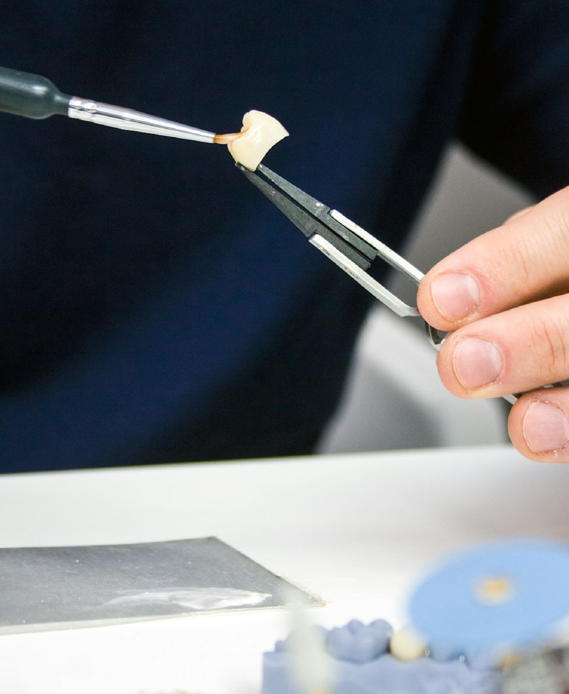 Hand holding tweezers with dental crown being painted with a brush over a workbench.