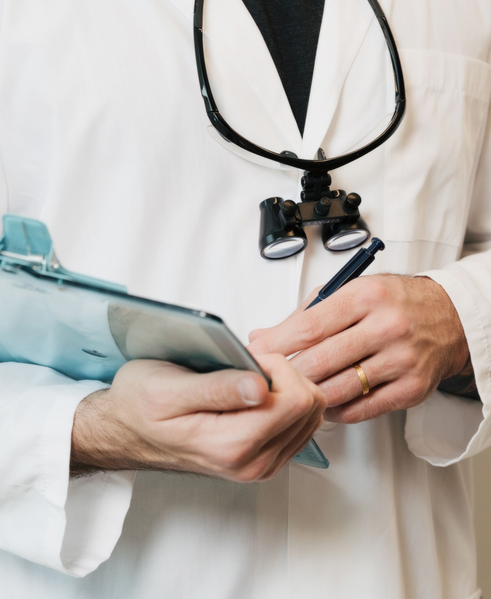 Doctor in white coat holding clipboard and pen with magnifying loupes around neck.
