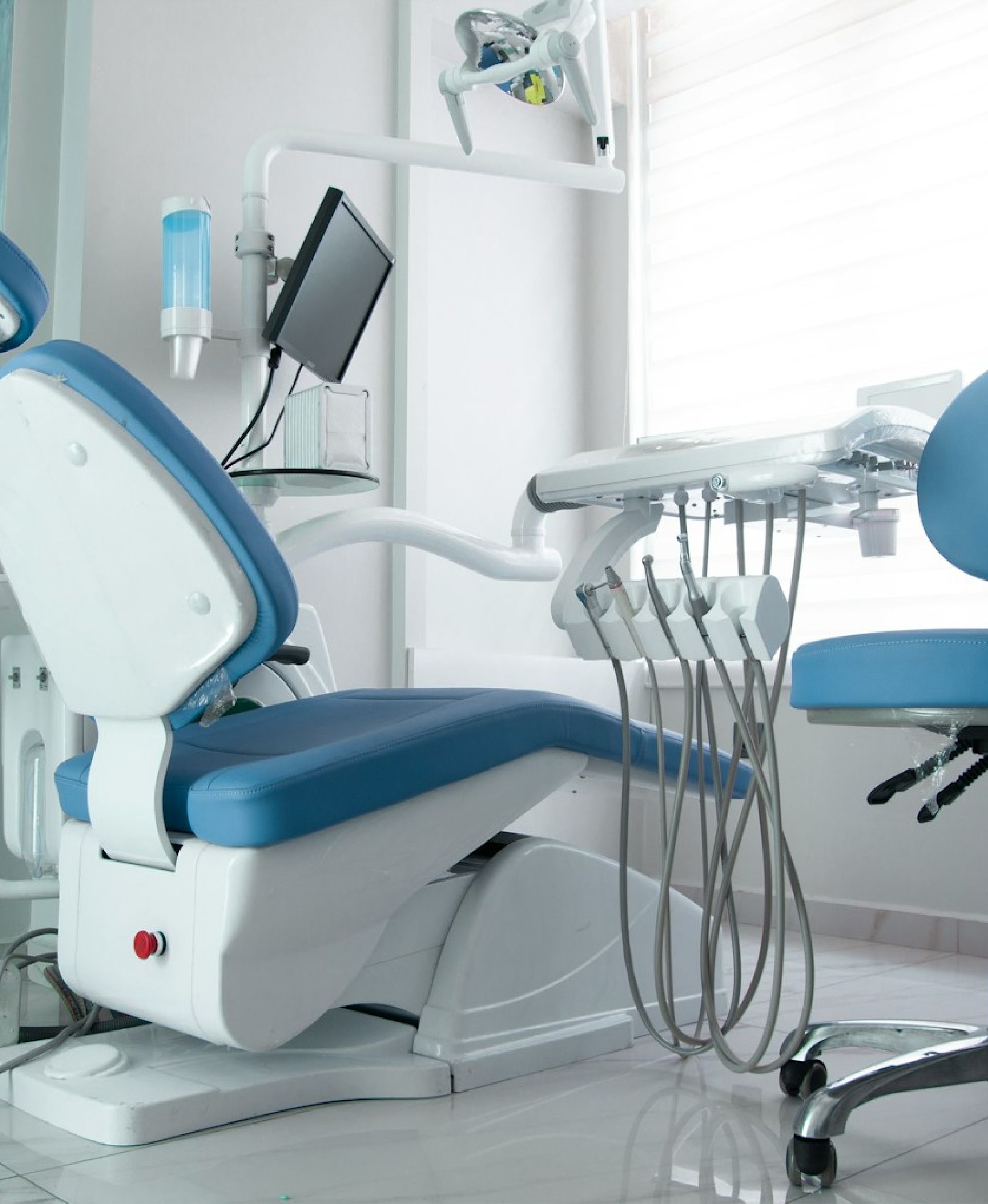 Empty dental chair with attached dental instruments and a monitor in a clean clinic room.