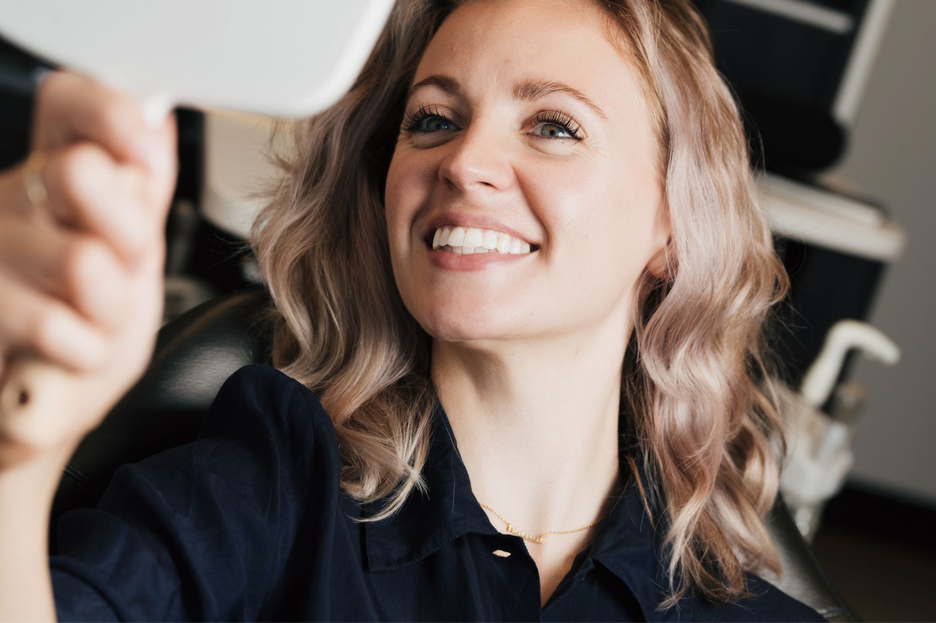 Smiling woman with wavy blonde hair looking at herself in a hand mirror.