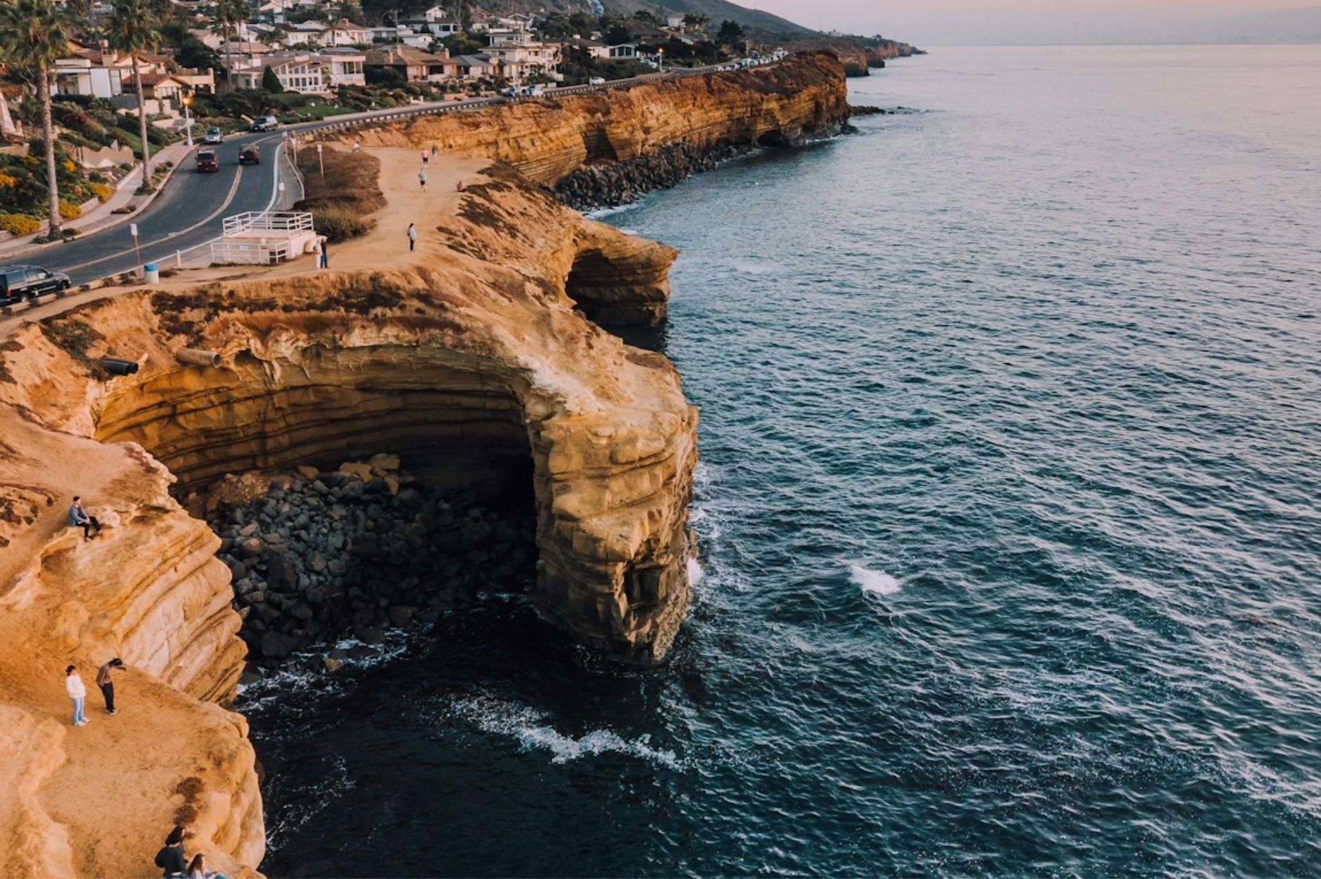 Ocean waves hitting rocky cliffs with people walking and sitting along the coastal path.