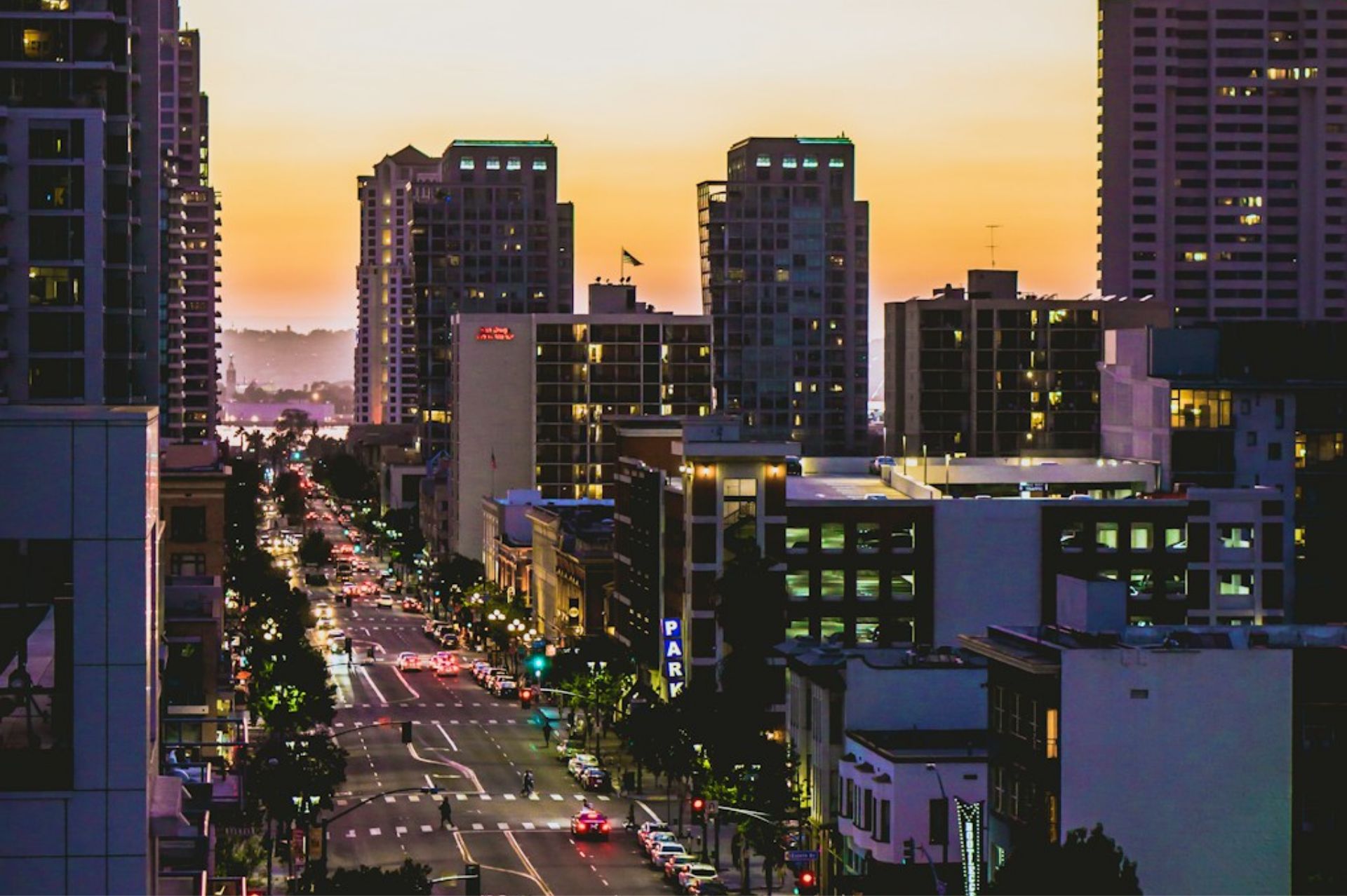 City street with traffic and tall buildings at sunset with orange sky in the background.