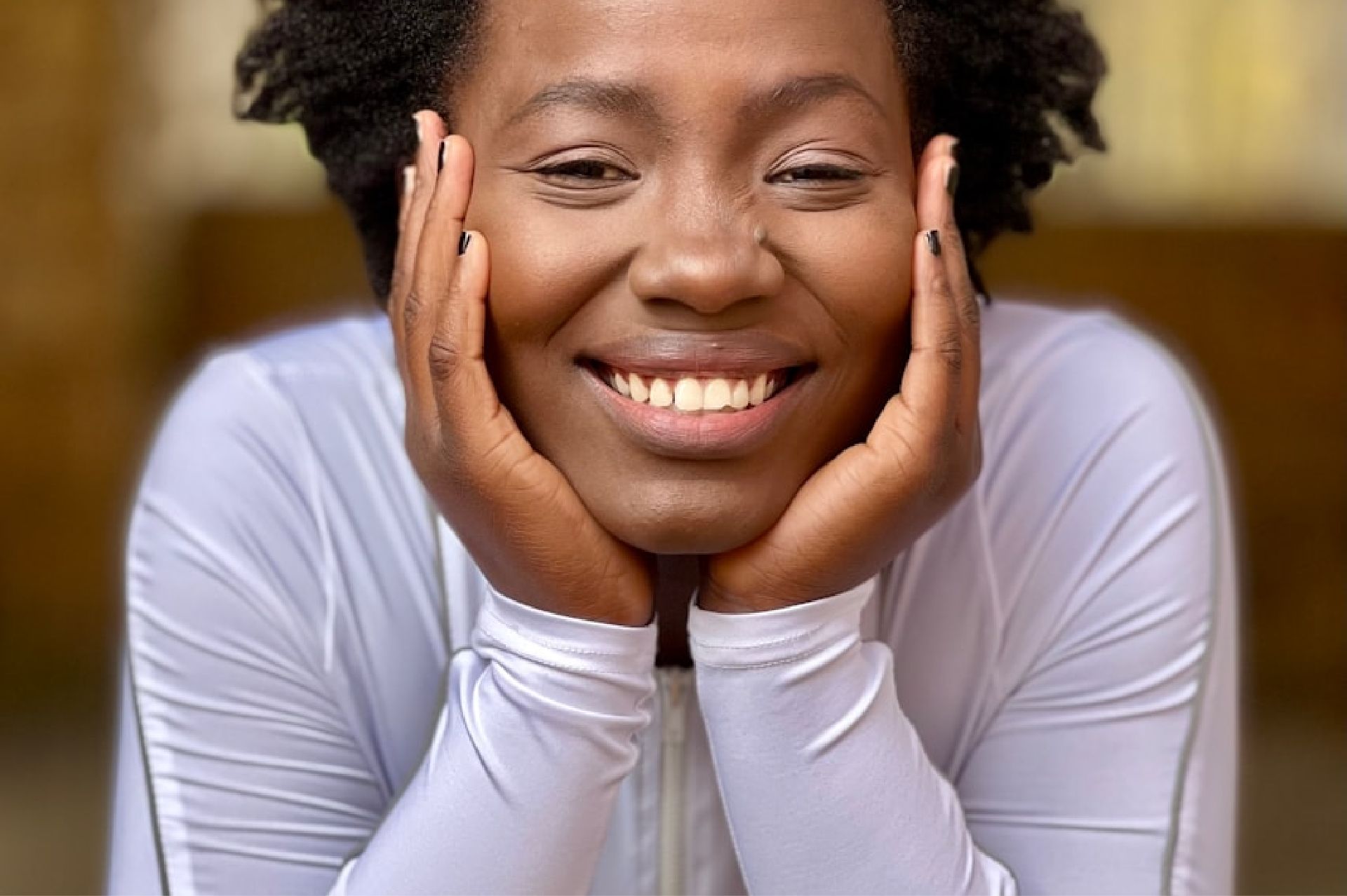 Close-up of a smiling woman resting her face on her hands wearing a white long-sleeve top.