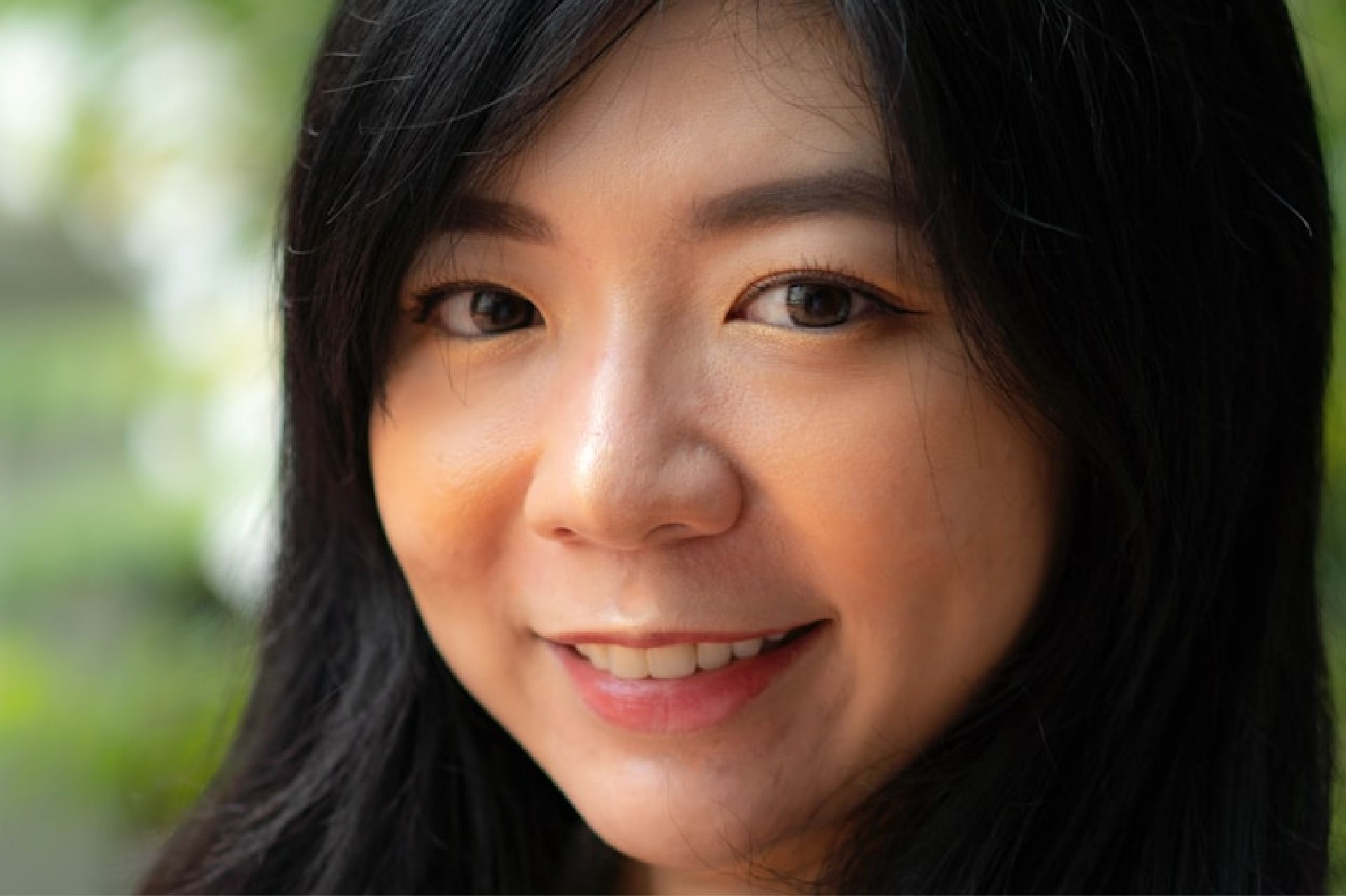 Close-up of a smiling woman with long black hair and natural makeup in a blurred outdoor setting.
