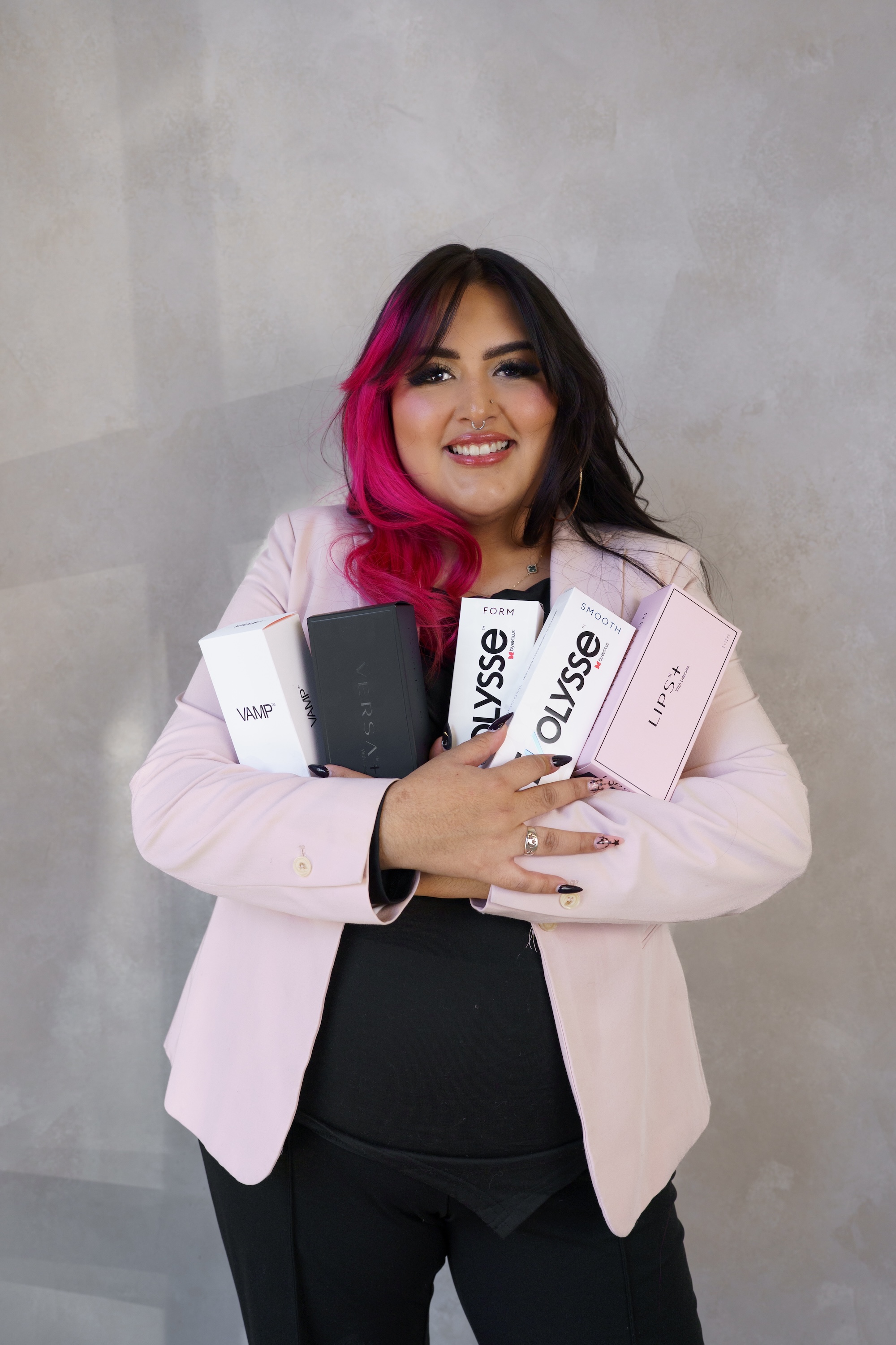 Smiling woman with half pink hair holding beauty product boxes in front of a gray wall.